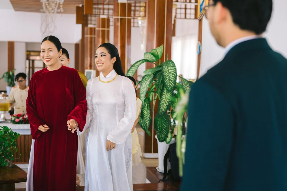 A bride in white holding hands with a woman in red, facing a groom