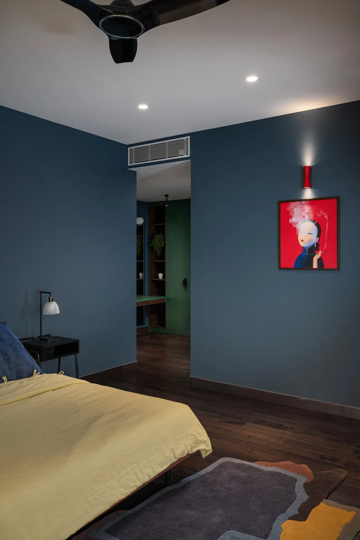 A bedroom with dark blue walls, a yellow bedspread, a black nightstand with a white lamp, and a framed artwork of a woman smoking. There's an open doorway leading to a room with green doors and wooden shelving, with hardwood floors and a colorful geometric rug.
