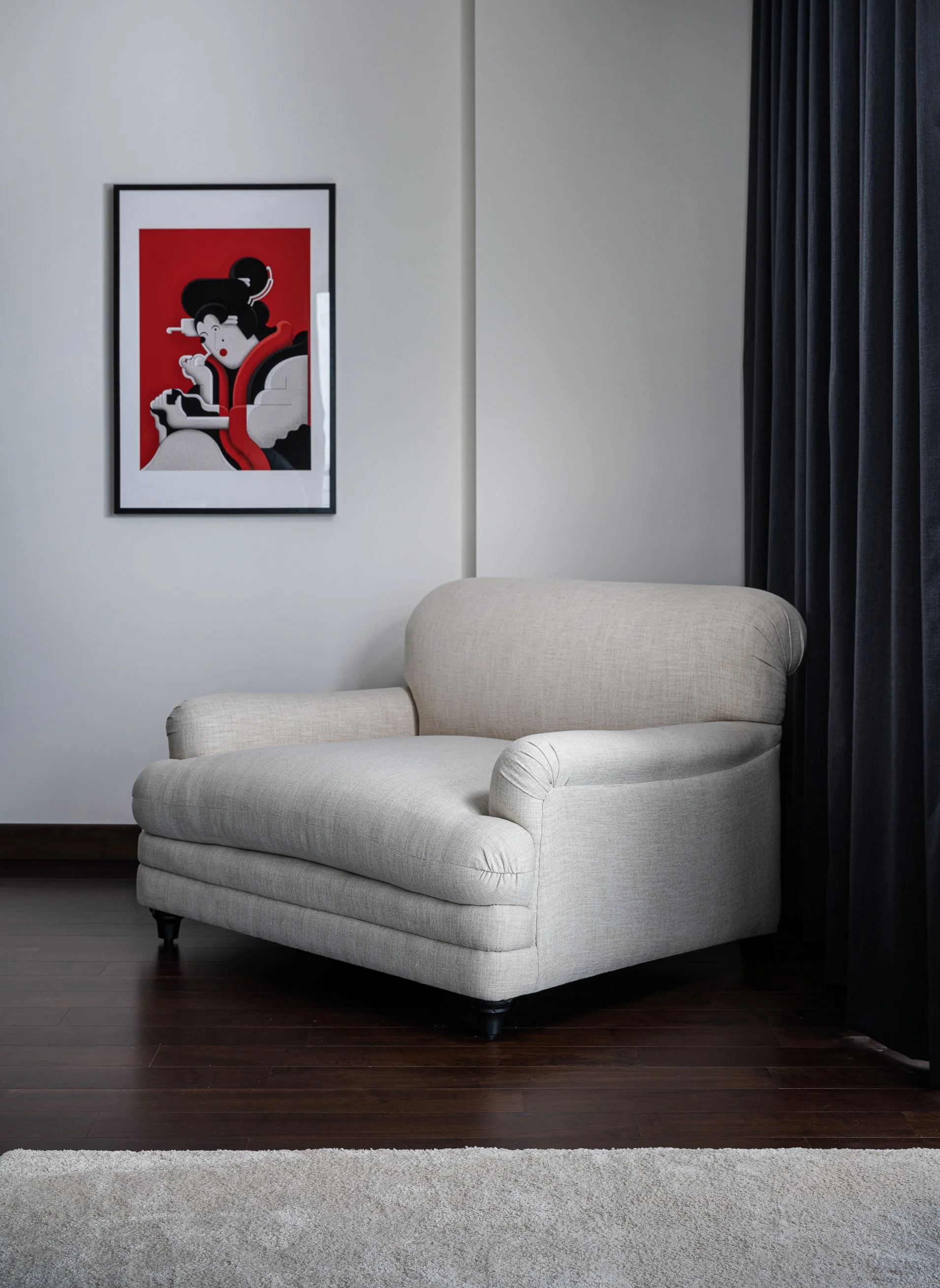 A beige armchair in a living room with dark wood flooring, a white wall with a framed artwork of a woman with black hair and red background, black curtains on the right, and a light-colored rug in front of the chair.