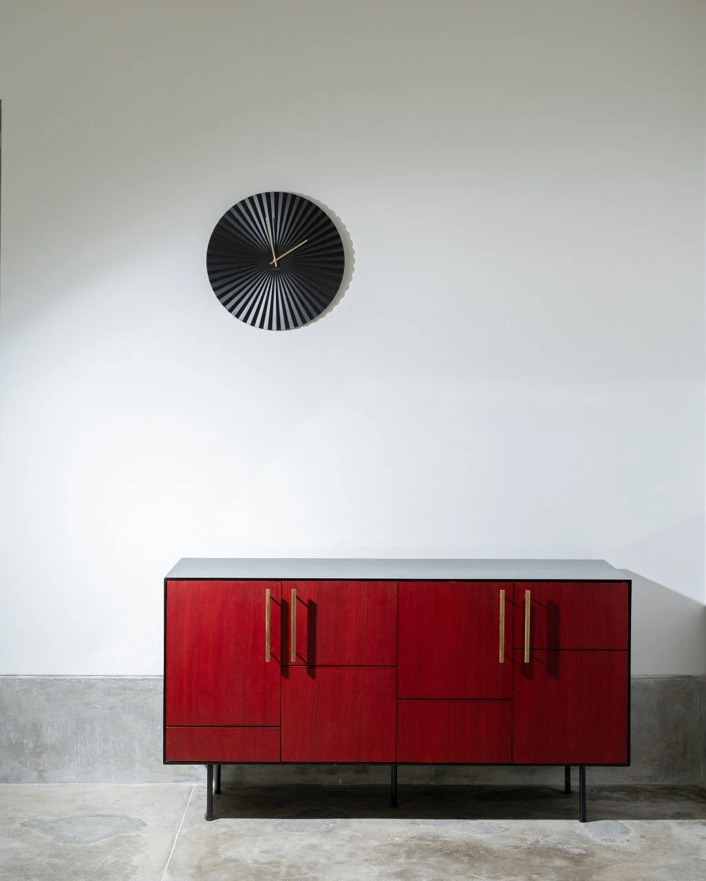 Minimalist interior with a white wall, a black circular wall clock with gold hands, and a modern red cabinet with gold handles on a concrete floor.