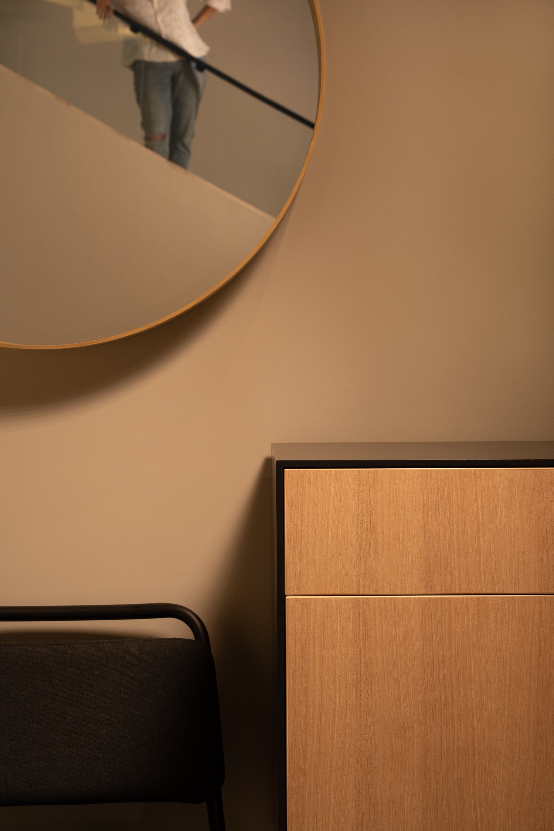 Part of a room with beige walls, a large round mirror reflecting a person in distressed jeans and a beige sweater, a black chair, and a wooden cabinet.