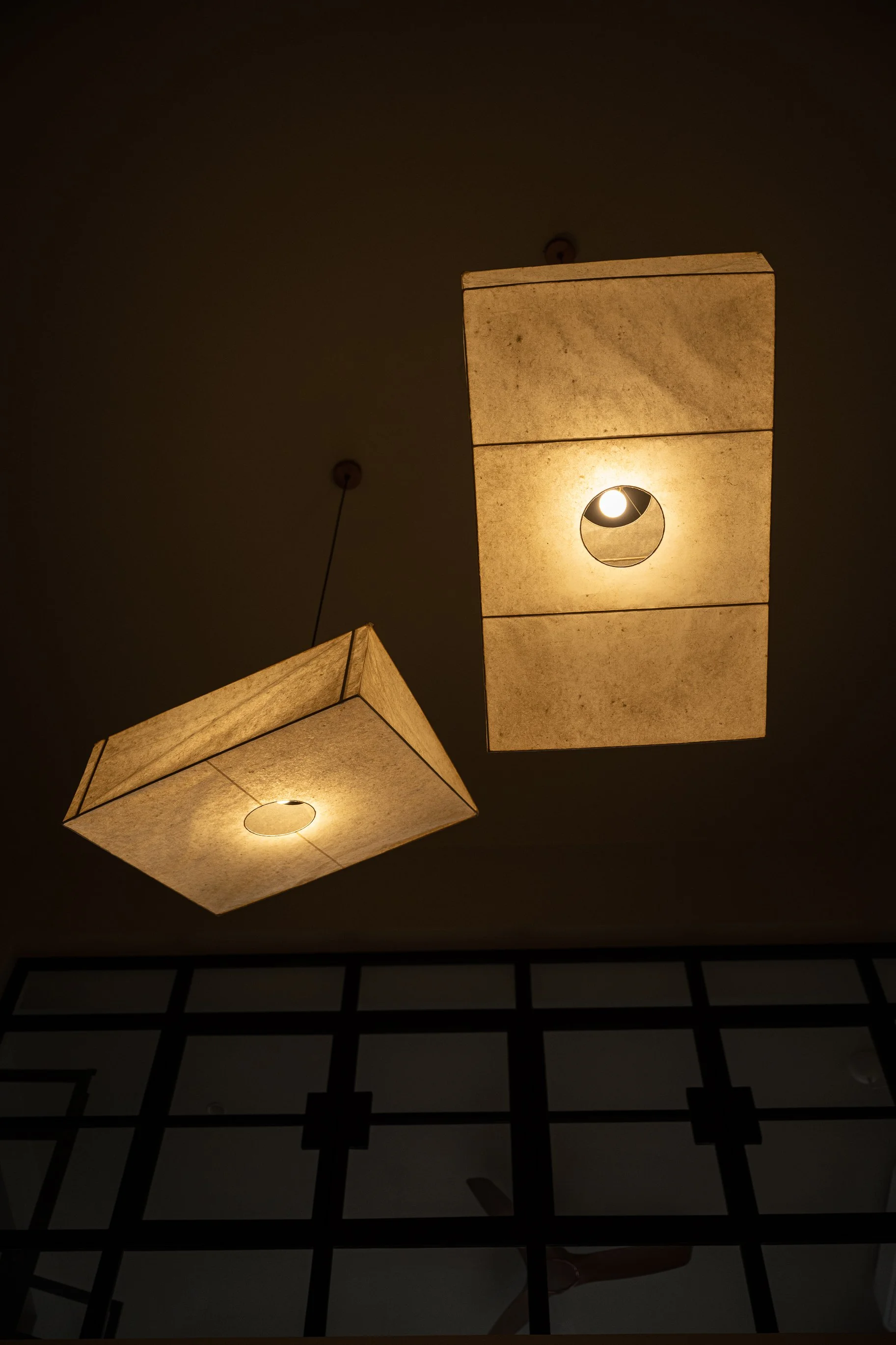 Two hanging ceiling lights with rectangular beige fabric shades and circular cutouts illuminating a dark ceiling, with black and white tiled floor below.