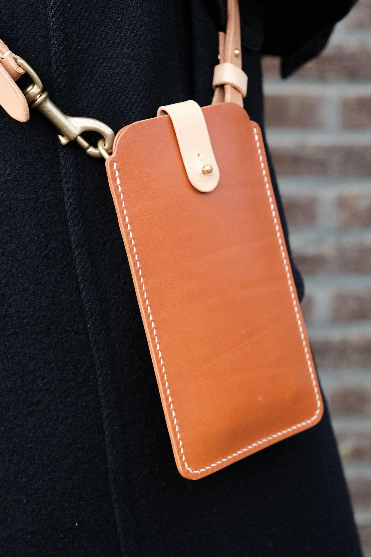 Close-up of a person wearing a black outfit carrying a tan and cream-colored leather phone holder with a clip and a strap, attached to a tan-colored bag with gold hardware.