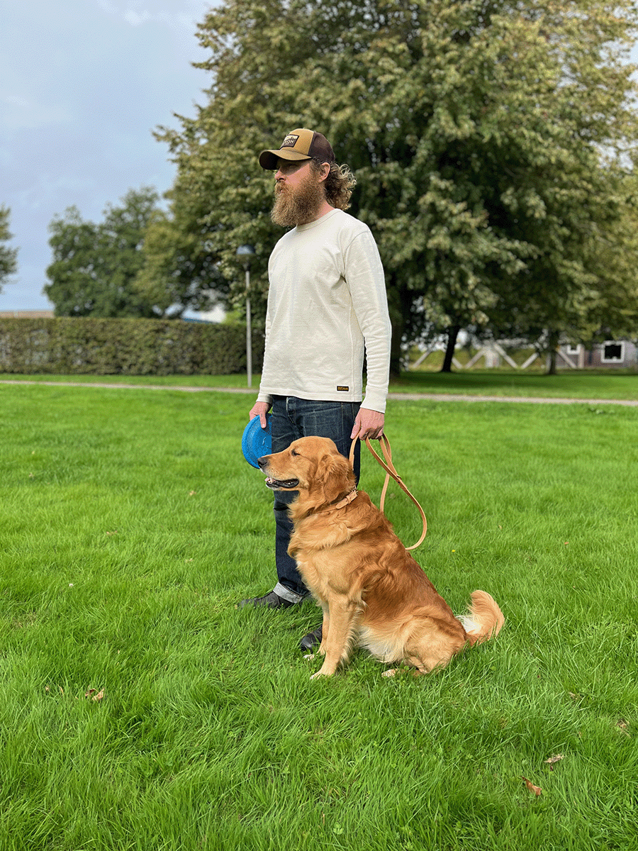 A man with a beard and wearing a white long-sleeve shirt, jeans, and a baseball cap stands on a green lawn with a golden retriever sitting beside him. The man holds a blue frisbee in one hand and a leash attached to the dog in the other. There are trees and a hedge in the background.