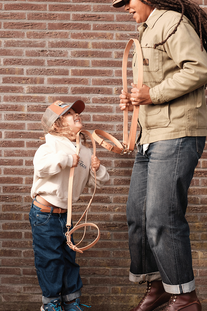 A woman and a young girl holding leashes of a dog, looking at each other with smiles against a brick wall background.