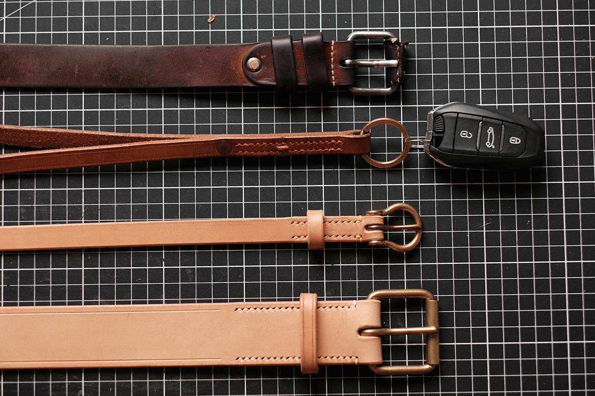 Four leather belts in various shades of brown, lined up horizontally, with a car key fob on top, all placed on a black grid background.
