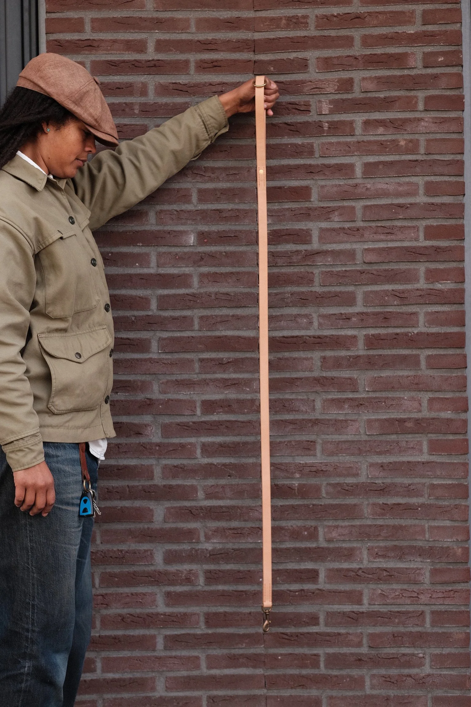 A woman wearing a beige jacket and a brown cap is standing next to a brick wall, holding a long, vertical measuring tape against it.