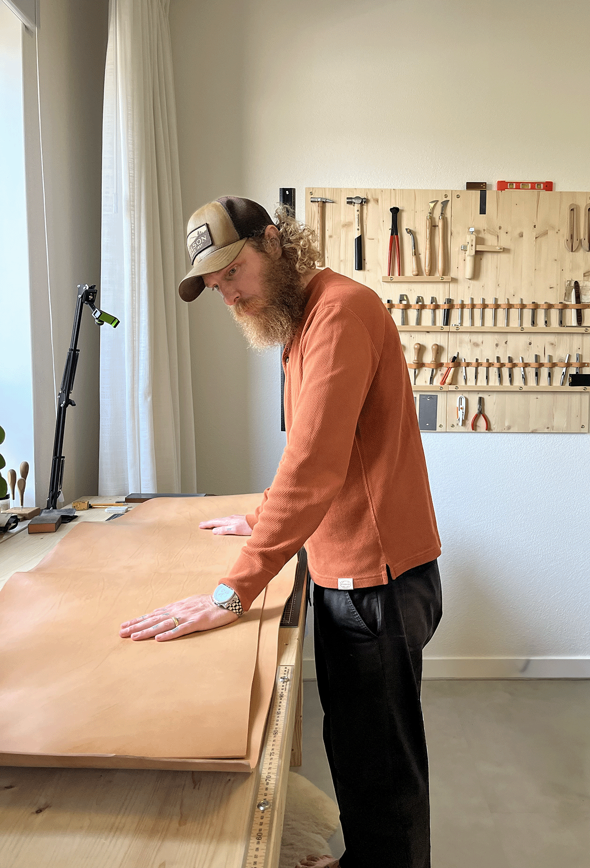 A man with a curly beard and long curly hair, wearing a trucker hat, orange shirt, and black pants, is standing at a workbench in a woodworking shop, pressing down on a large piece of leather or similar material on the table.