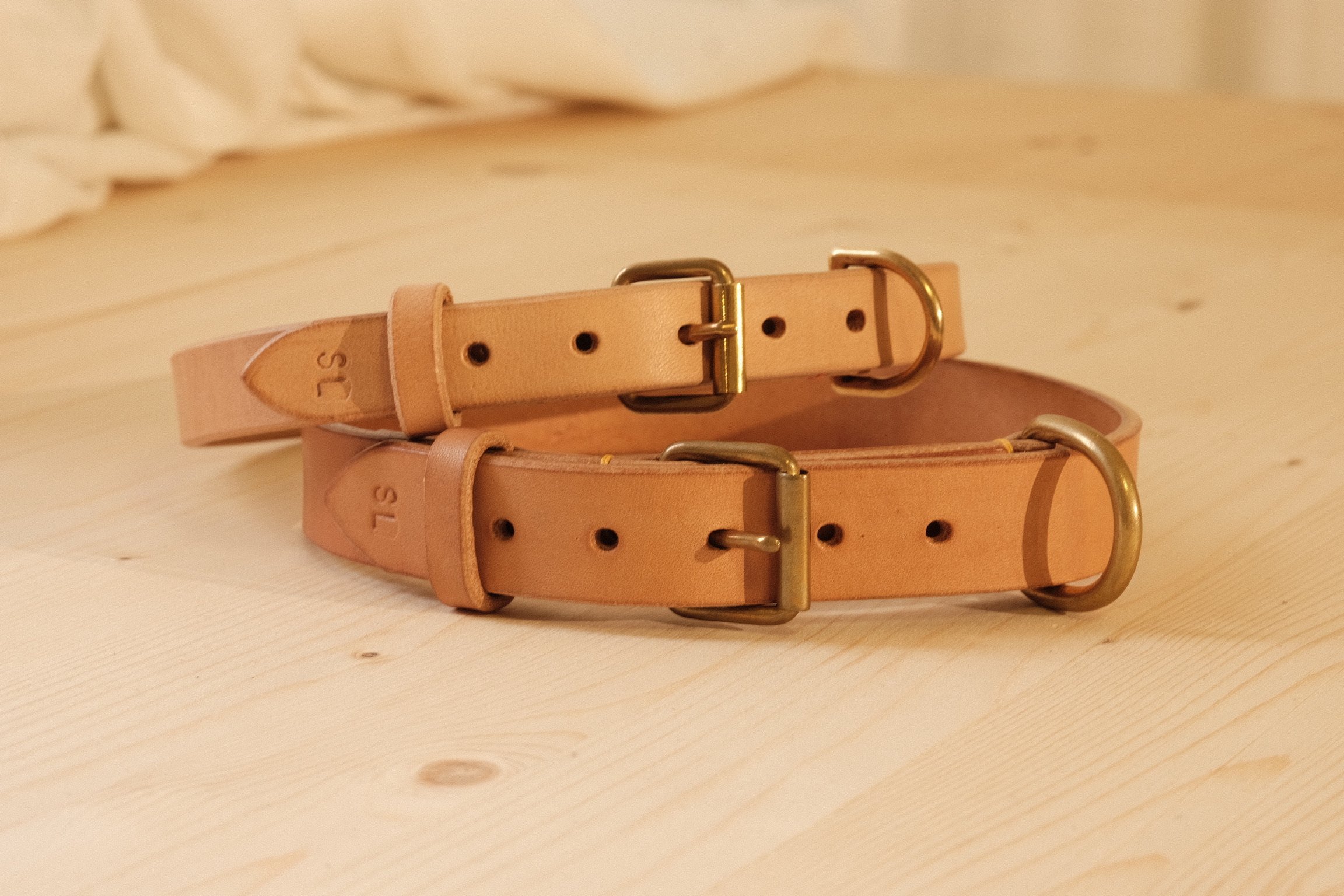 Two beige leather belts with brass buckles lying on a light wooden surface.
