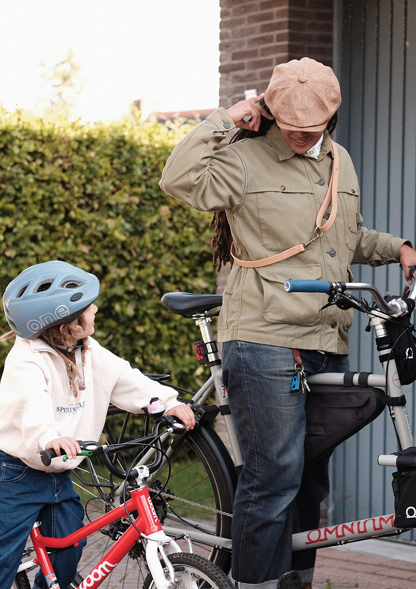 A woman with the crossbody Heritage Leather Lead from S T U D I O  V A N  D E  L A A R next to a girl with a bike helmet, both with bikes, standing outside near a brick building and bushes.