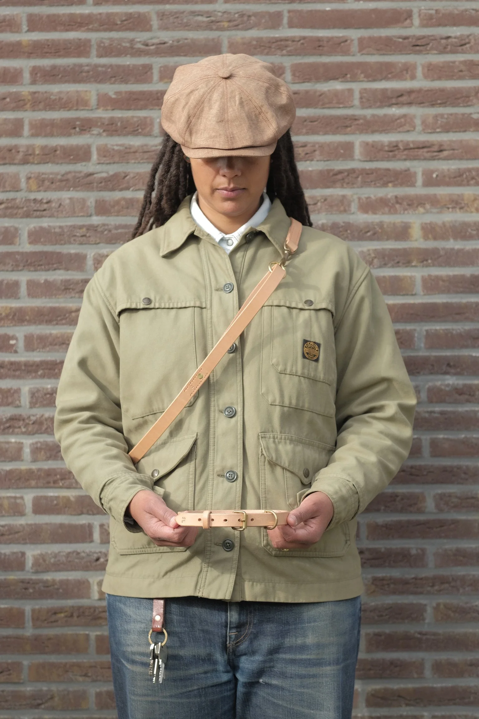Person wearing a beige cap, beige jacket, and jeans, standing against a brick wall, holding a light pink belt with gold buckle and keys attached.