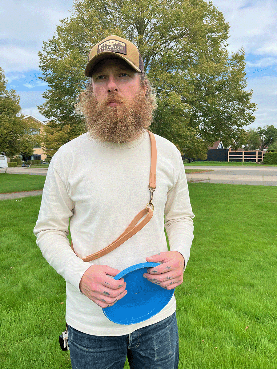 A man with a beard and tattoos on his fingers stands outdoors and is playing with his dog.  He's wearing a beige long-sleeve shirt, a brown cap, and has the Leather Heritage Lead from S T U D I O  V A N  D E  L A AR  crossbody over his shoulder.
