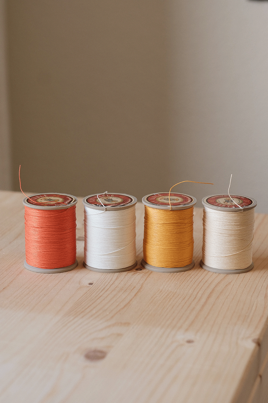 Four spools of thread in red, white, gold, and cream colors, arranged in a row on a wooden surface.