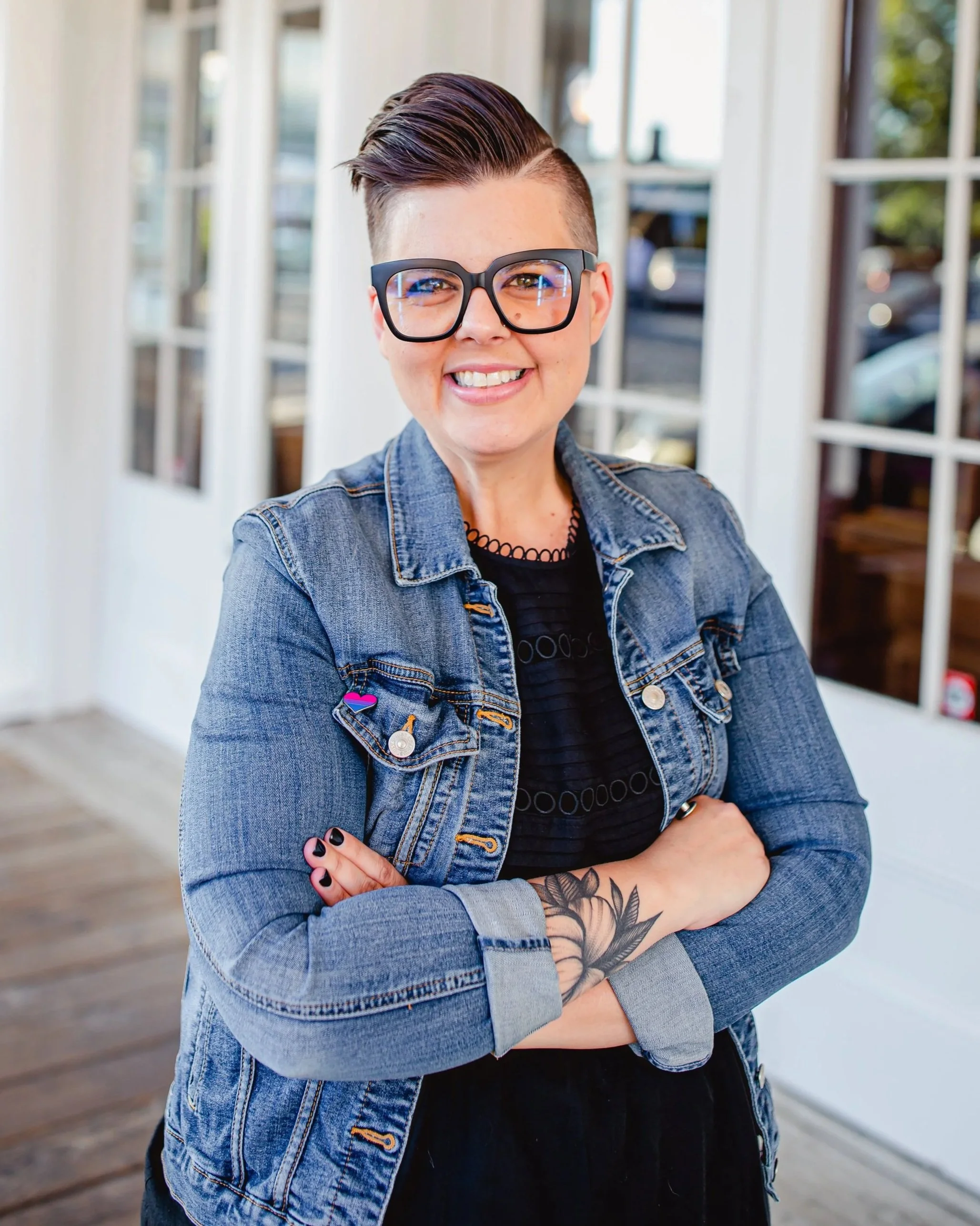 A person with short dark hair, glasses, and tattoos on their arms, smiling with arms crossed in front of a bright window and white walls.