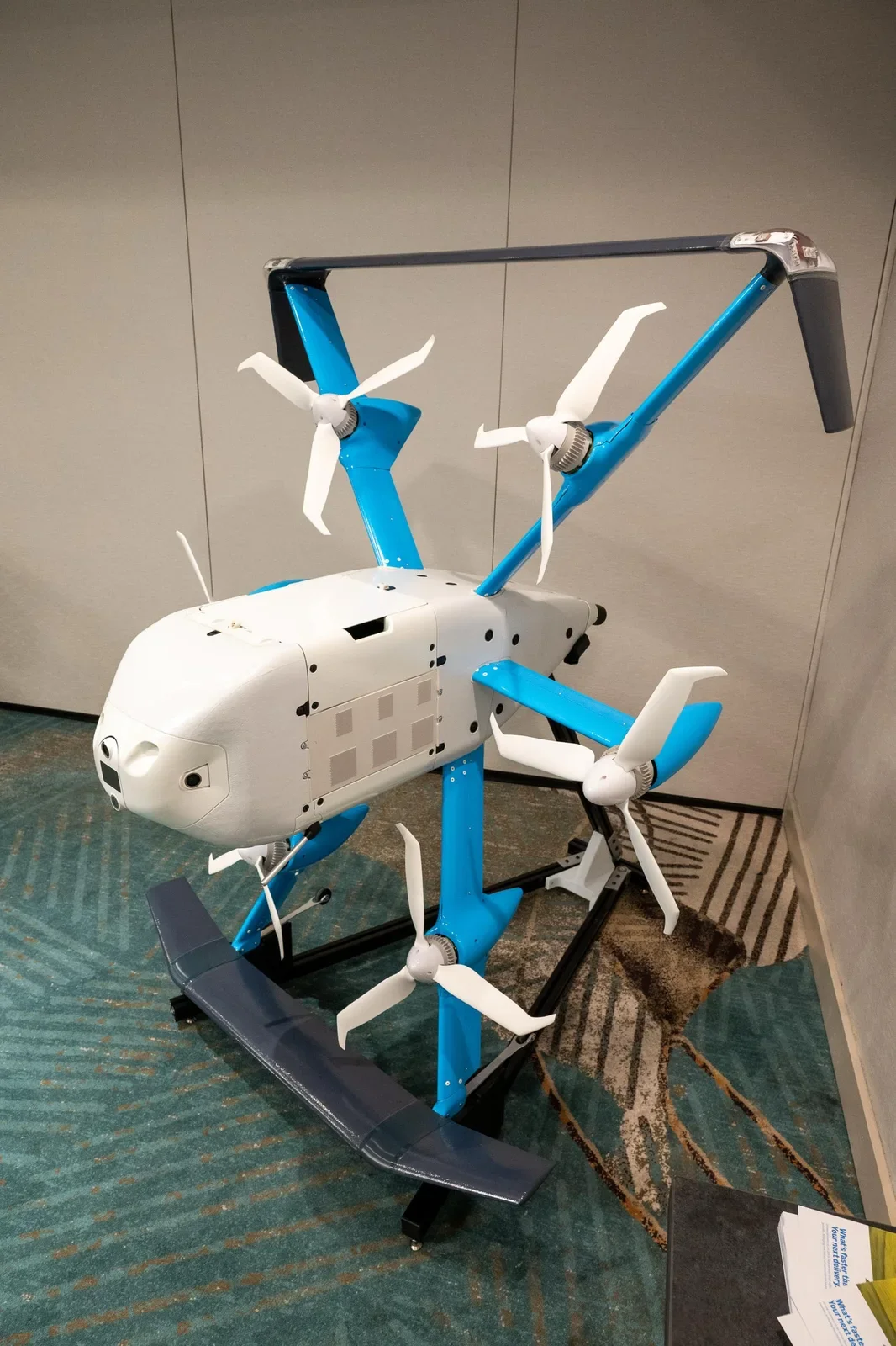 A robot with a helicopter shape, featuring multiple white and blue propellers and a white body, displayed indoors on a patterned carpeted floor.
