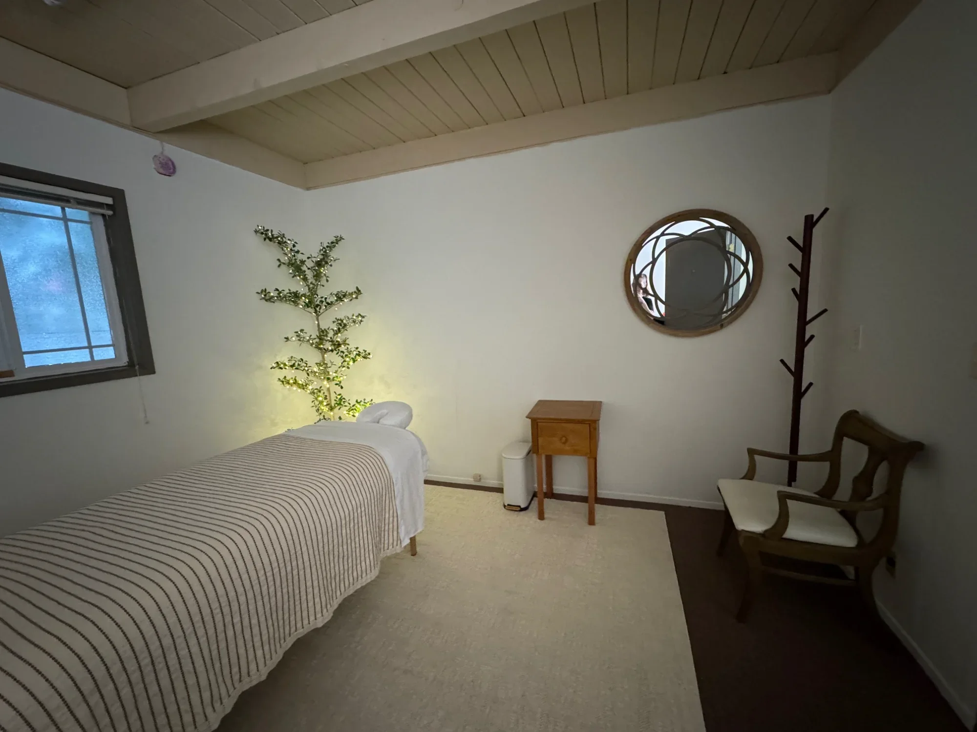A bedroom with two beds with striped bedspreads, a small wooden nightstand, a decorative round mirror on the wall, a wooden coat rack, a wooden chair with a cushion, a window with blinds, and a small potted plant in the corner with string lights.