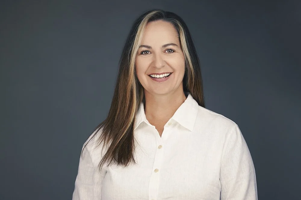 A woman with long brown hair and blonde highlights smiling at the camera, wearing a white button-up shirt, against a dark gray background.
