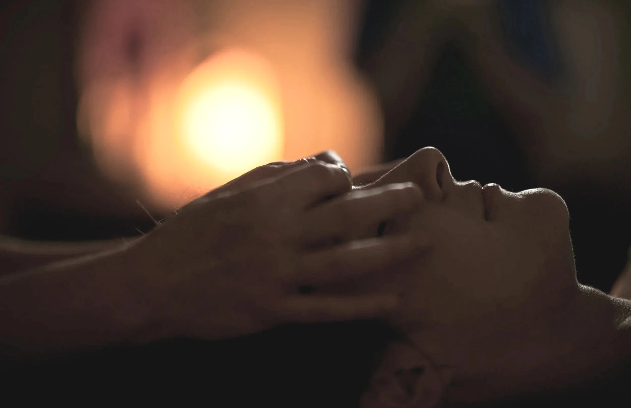 Close-up of a person's face lying down, with their hand gently resting on their forehead, illuminated by warm, soft lighting.