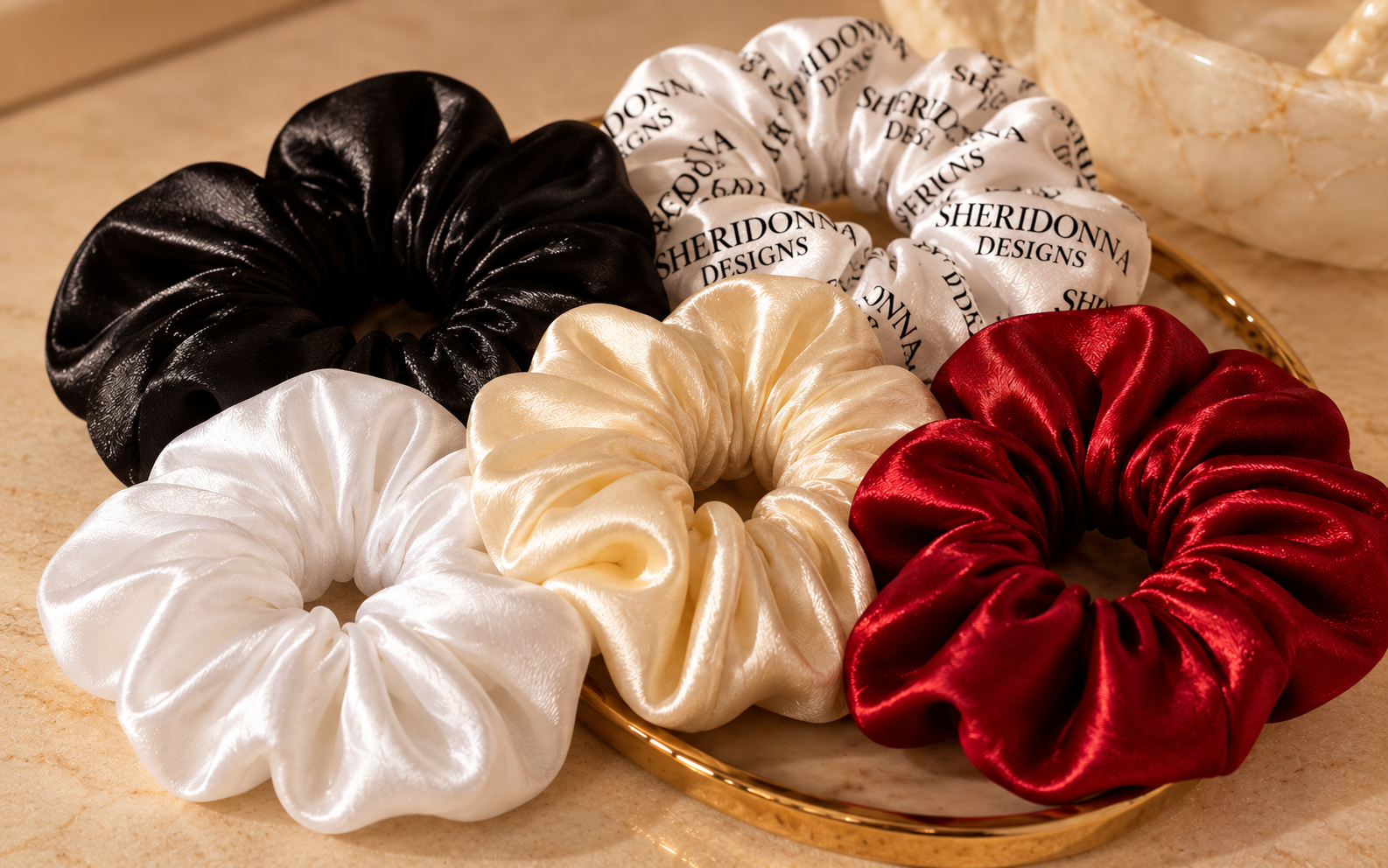 Five luxurious Sheridonna Designs satin scrunchies in Midnight Noir, Pure White, Ivory, Candy Apple Red and the signature black-on-white print, elegantly arranged on a gold-trimmed tray.