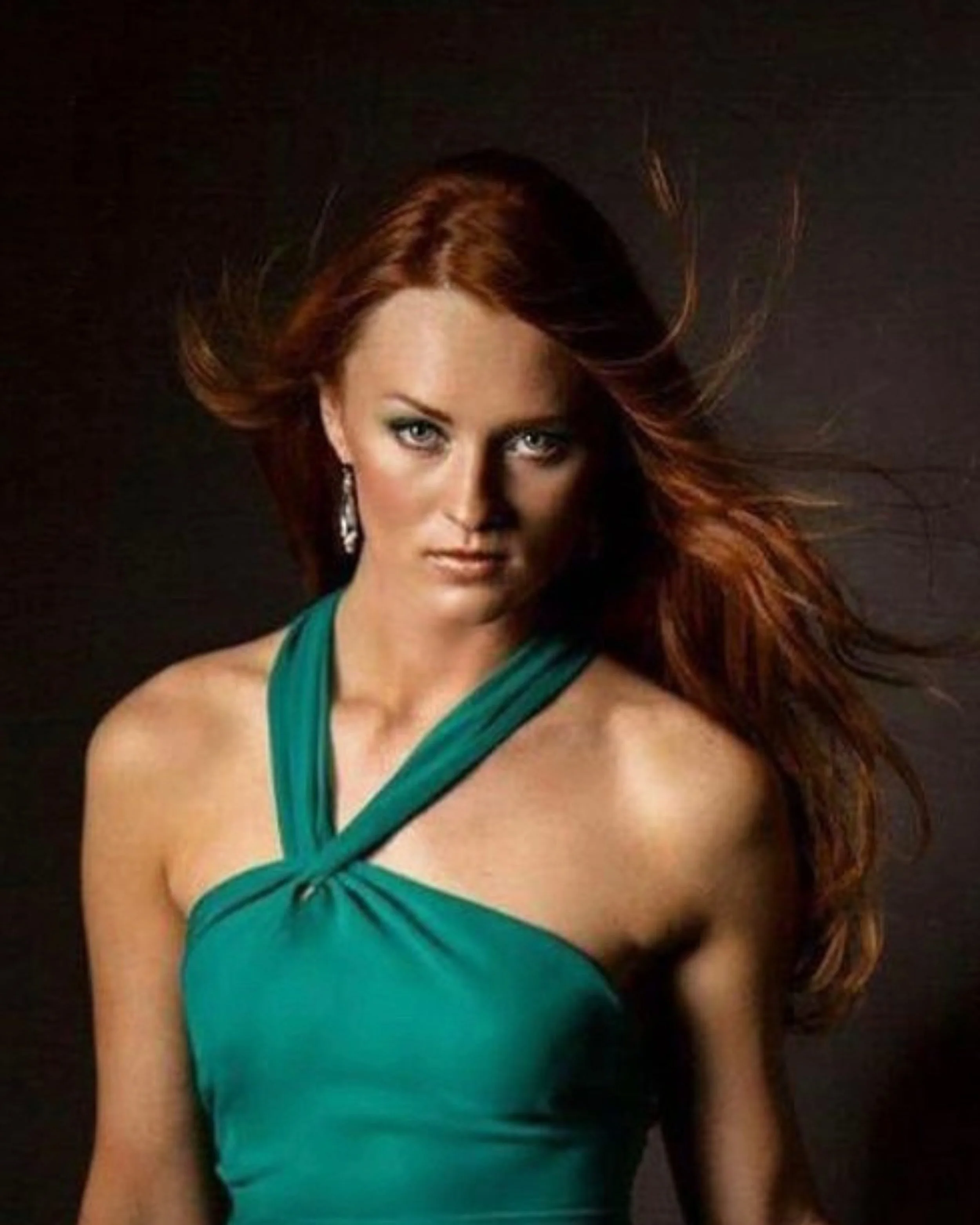 Woman with long, voluminous auburn hair styled in flowing waves, wearing a teal halter dress against a dark studio background.