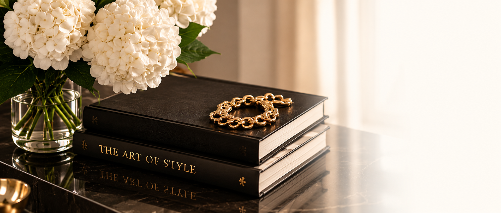 White hydrangeas beside stacked black fashion books and a gold bracelet on a marble table in a luxurious interior.