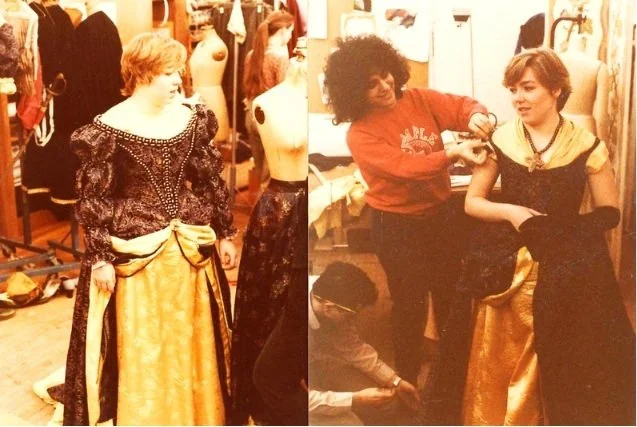 A woman with curly hair is helping a young woman try on a fancy dress in a costume shop. An older woman with short blond hair is standing nearby, observing. The shop has mannequins and costumes in the background.