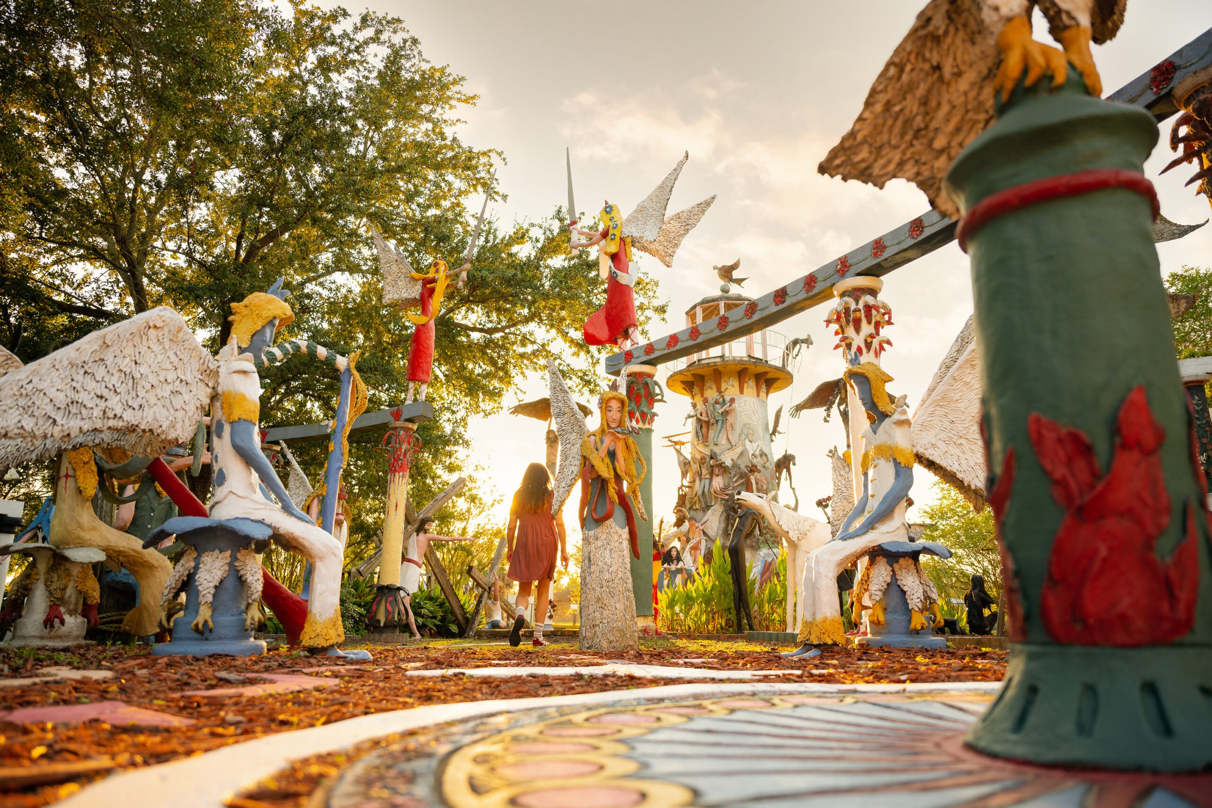 Colorful outdoor sculpture garden with fairy figurines and fairy tale characters, set against a background of green trees and a partly cloudy sky, illuminated by warm sunlight.