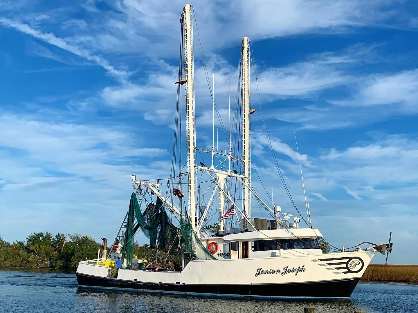 A white fishing boat named Jenson Joseph sailing on calm water with a blue sky, wispy clouds, and green shoreline in the background.