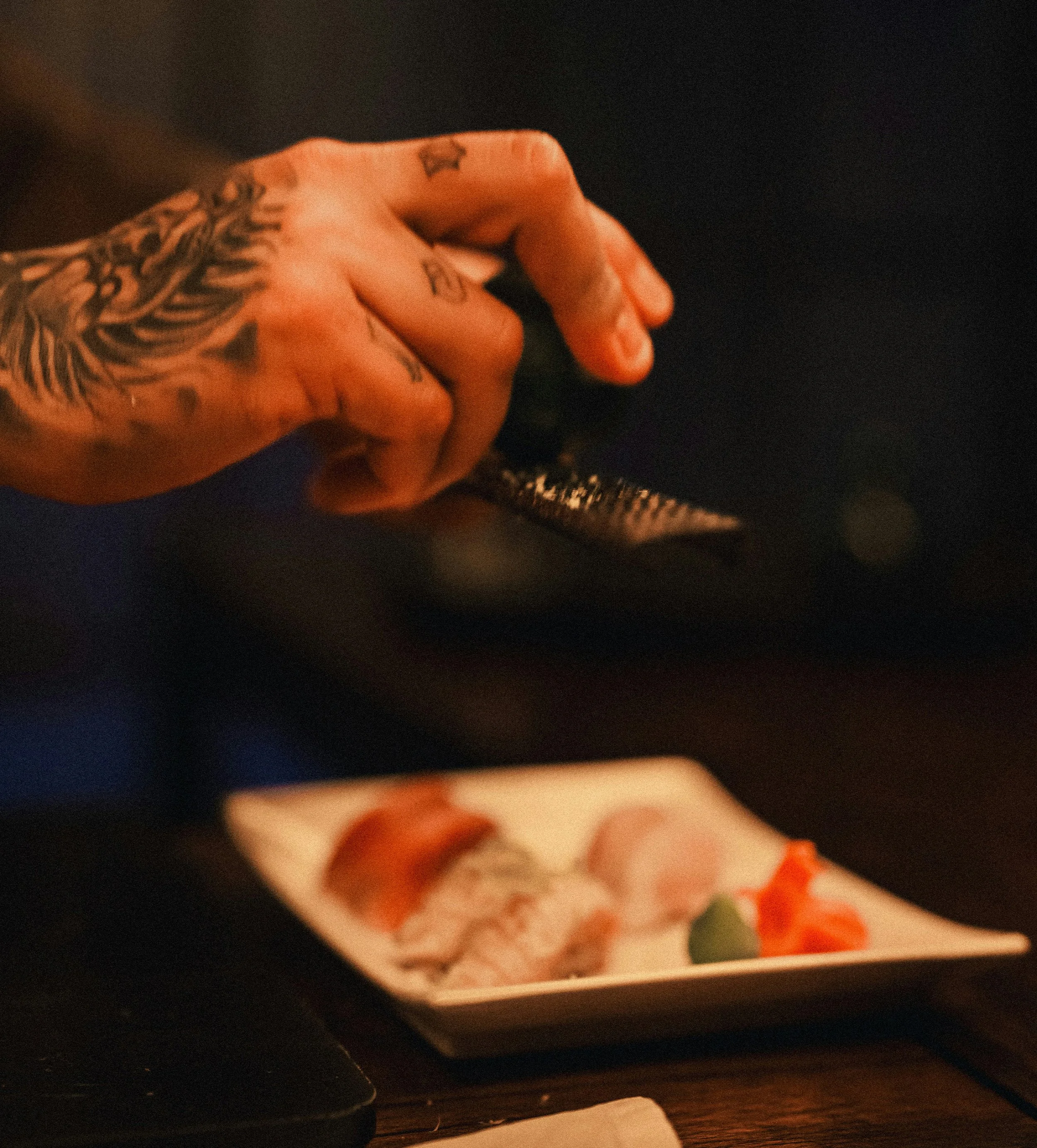 A close-up of a hand with tattoos, holding a piece of meat, possibly a slice of pizza, with some vegetables on it, against a dark background.