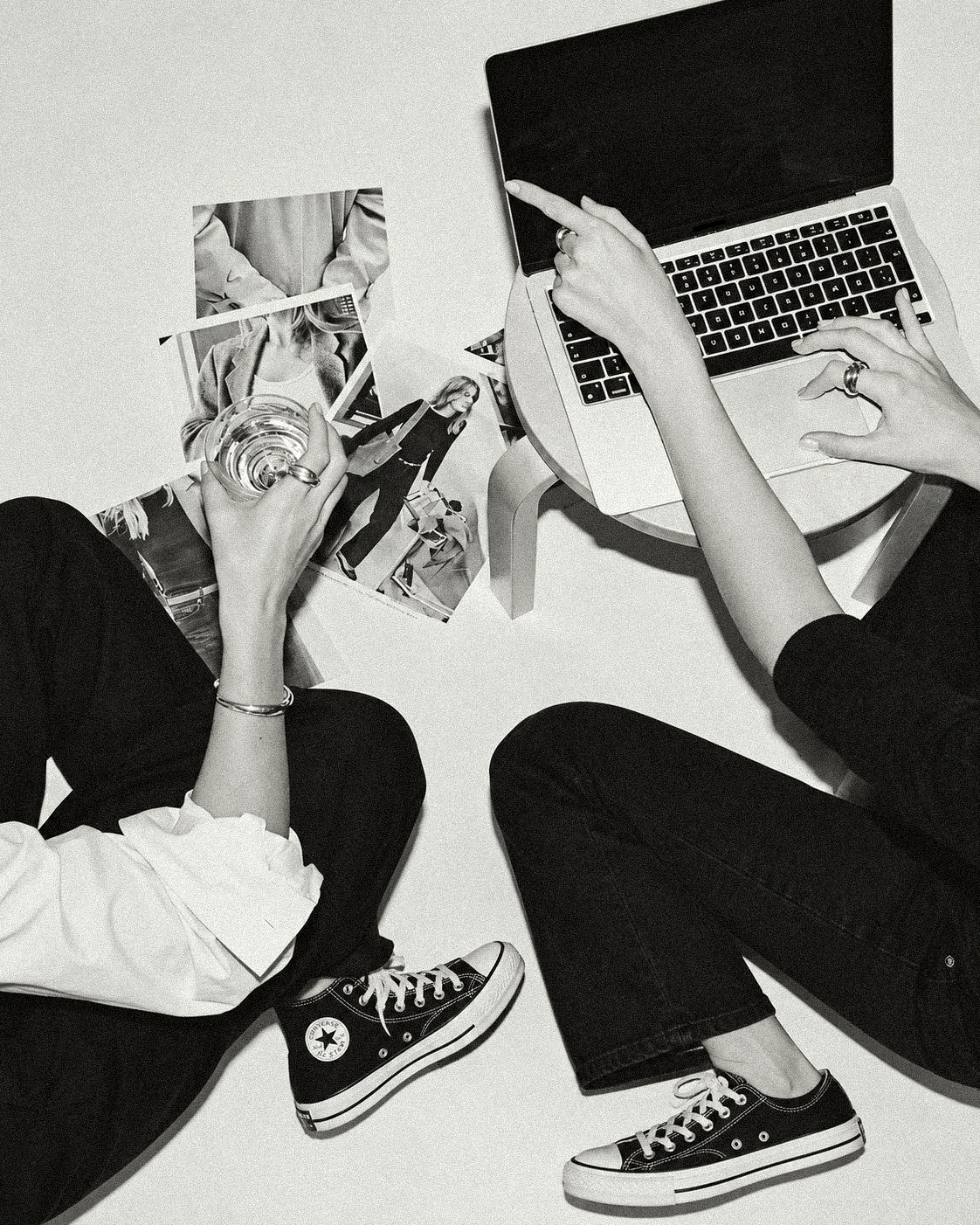 Two people sitting on the floor with a laptop, magazines, and a glass of water, wearing casual clothes and sneakers.