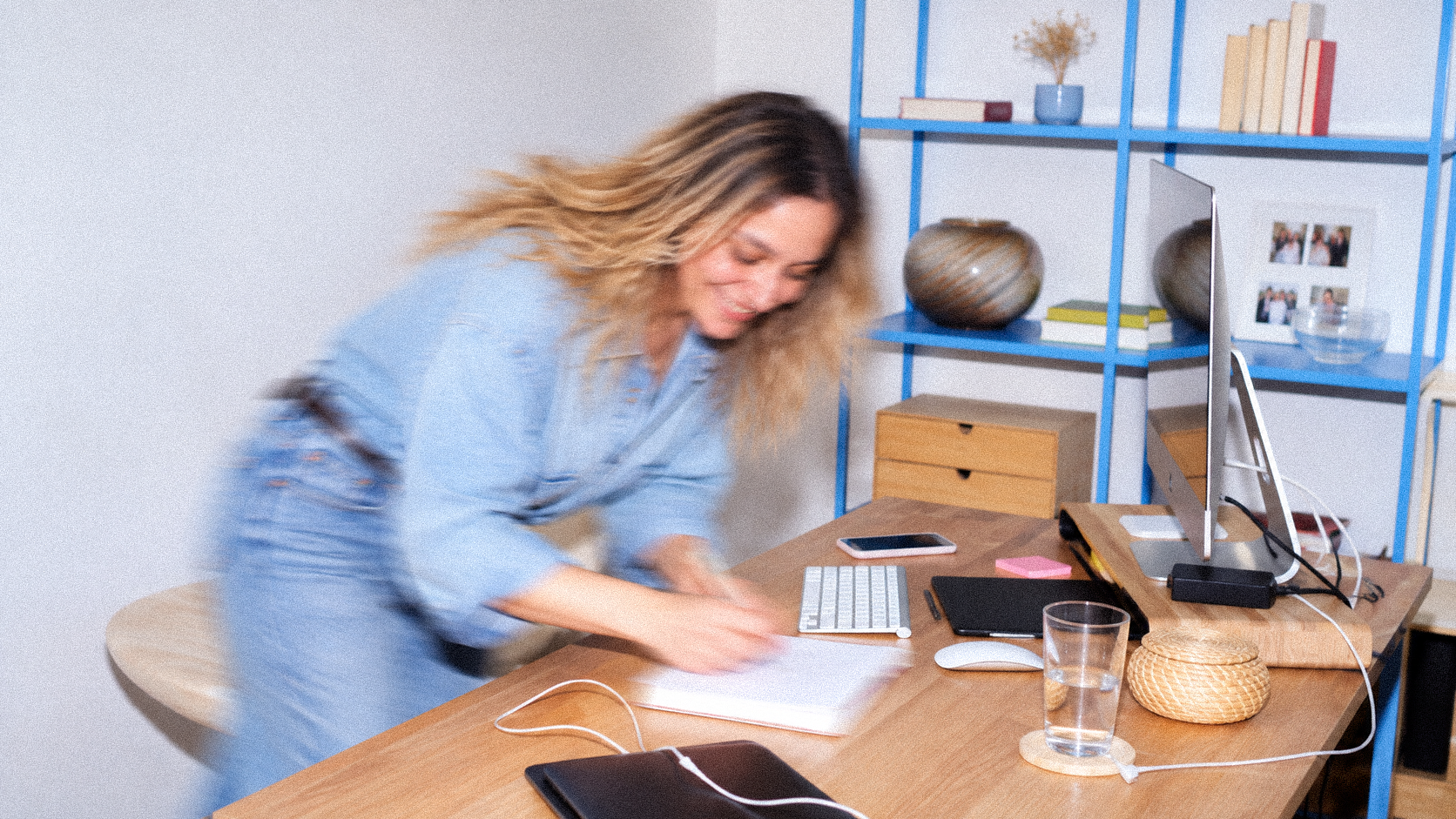 Woman with blonde, wavy hair smiling and leaning over a wooden desk, which has a computer, keyboard, smartphone, sticky notes, glass of water, and electronic accessories, in a modern office space with blue shelving and decorative items in the background.