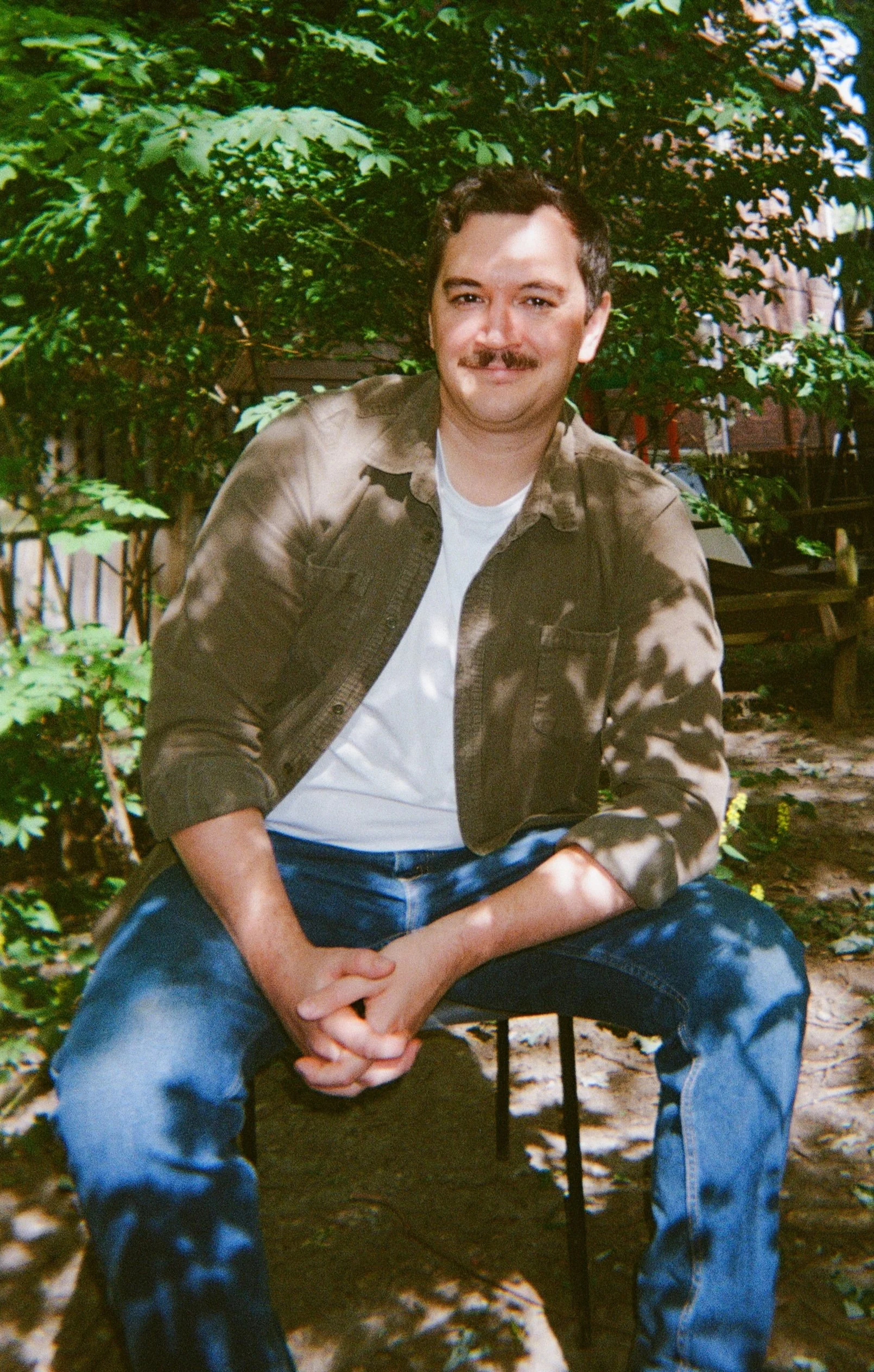A young man with a mustache, wearing a camouflage jacket over a white T-shirt and blue jeans, sitting outdoors in front of green foliage and trees, with dappled sunlight on his face.