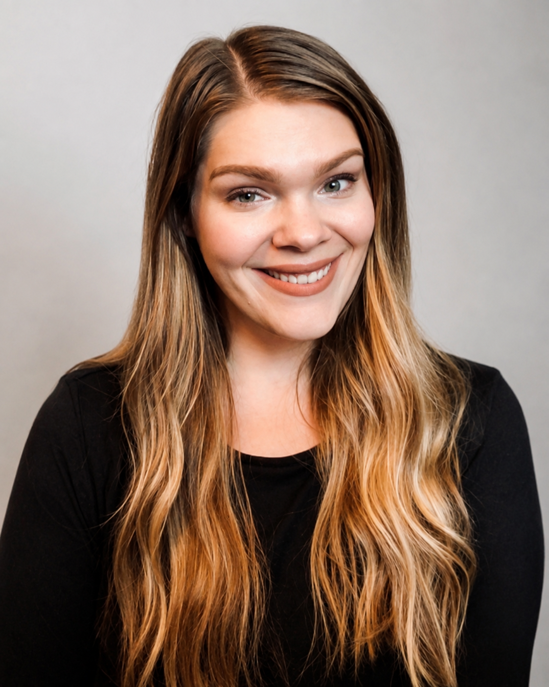 Portrait of a woman with long wavy light brown hair, fair skin, wearing a black top, smiling against a plain light gray background.