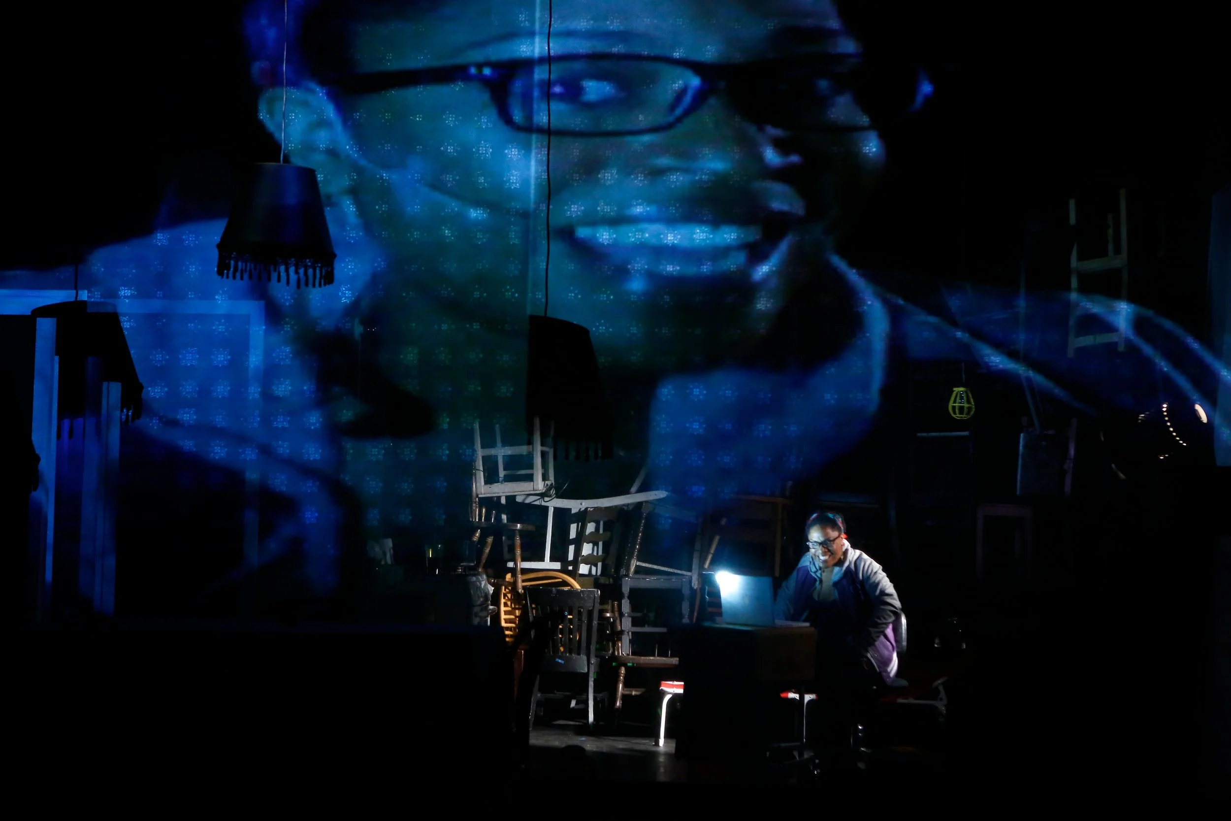 A large digital projection of a person's face with glasses on a wall, with a person working at a table in front of it, in a dark room.