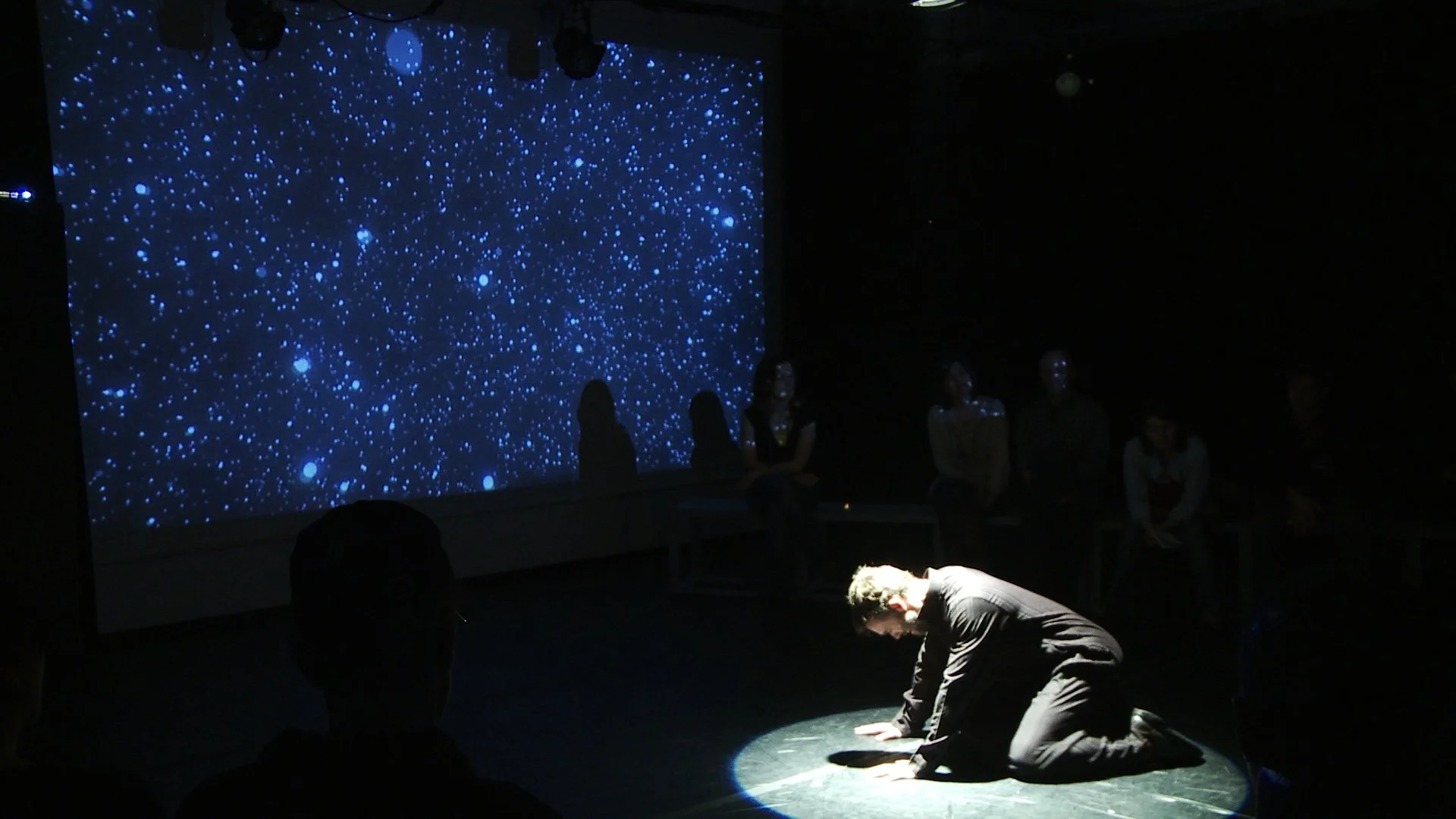 A person is bowing on the floor, illuminated by a spotlight, in front of an audience in a dimly lit room. A large screen behind displays a starry night sky.