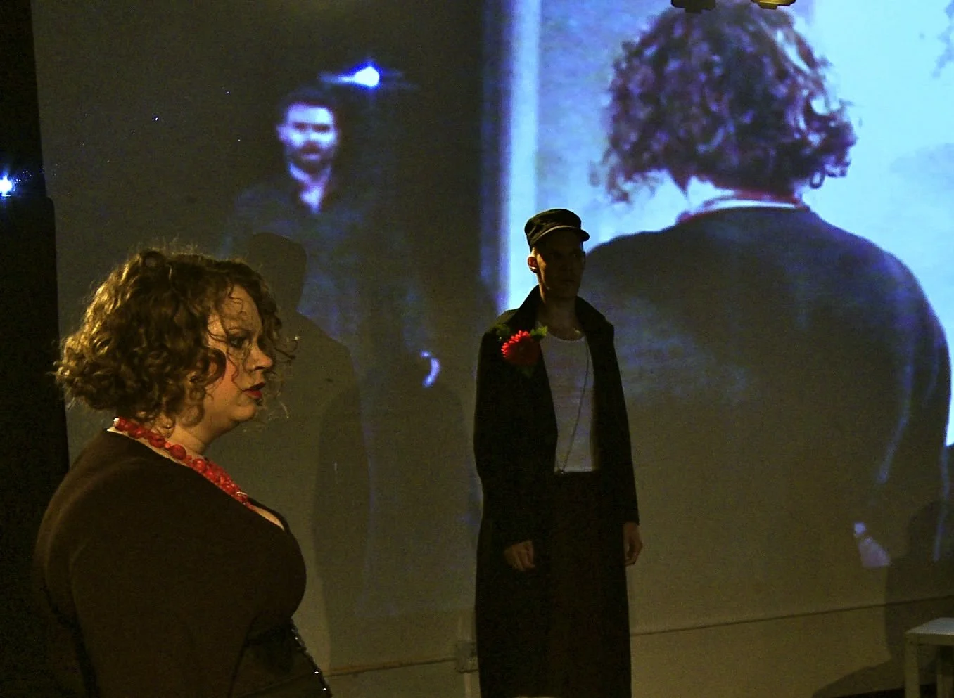 Three individuals standing in front of a projected black-and-white film or photograph, with one woman in the foreground looking downward, wearing a dark outfit and red necklace, and two men in the background, one of whom is wearing a hat and a flower on his chest.