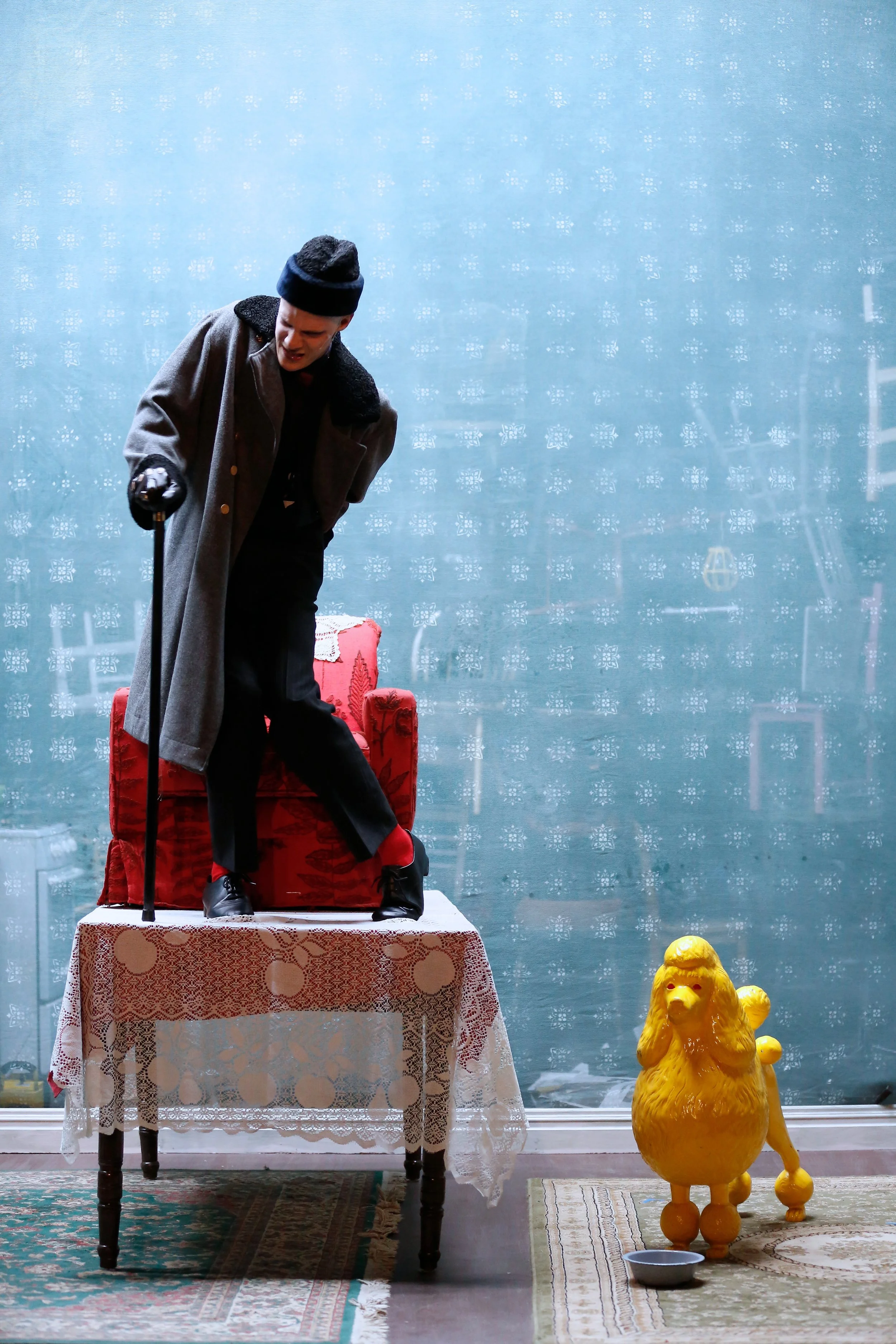 A person dressed in a long coat and black hat is standing on a table, leaning forward with a cane in hand, next to a red upholstered chair, with a yellow lion-shaped figure nearby, in front of a blue window with snowflake patterns and a green rug.