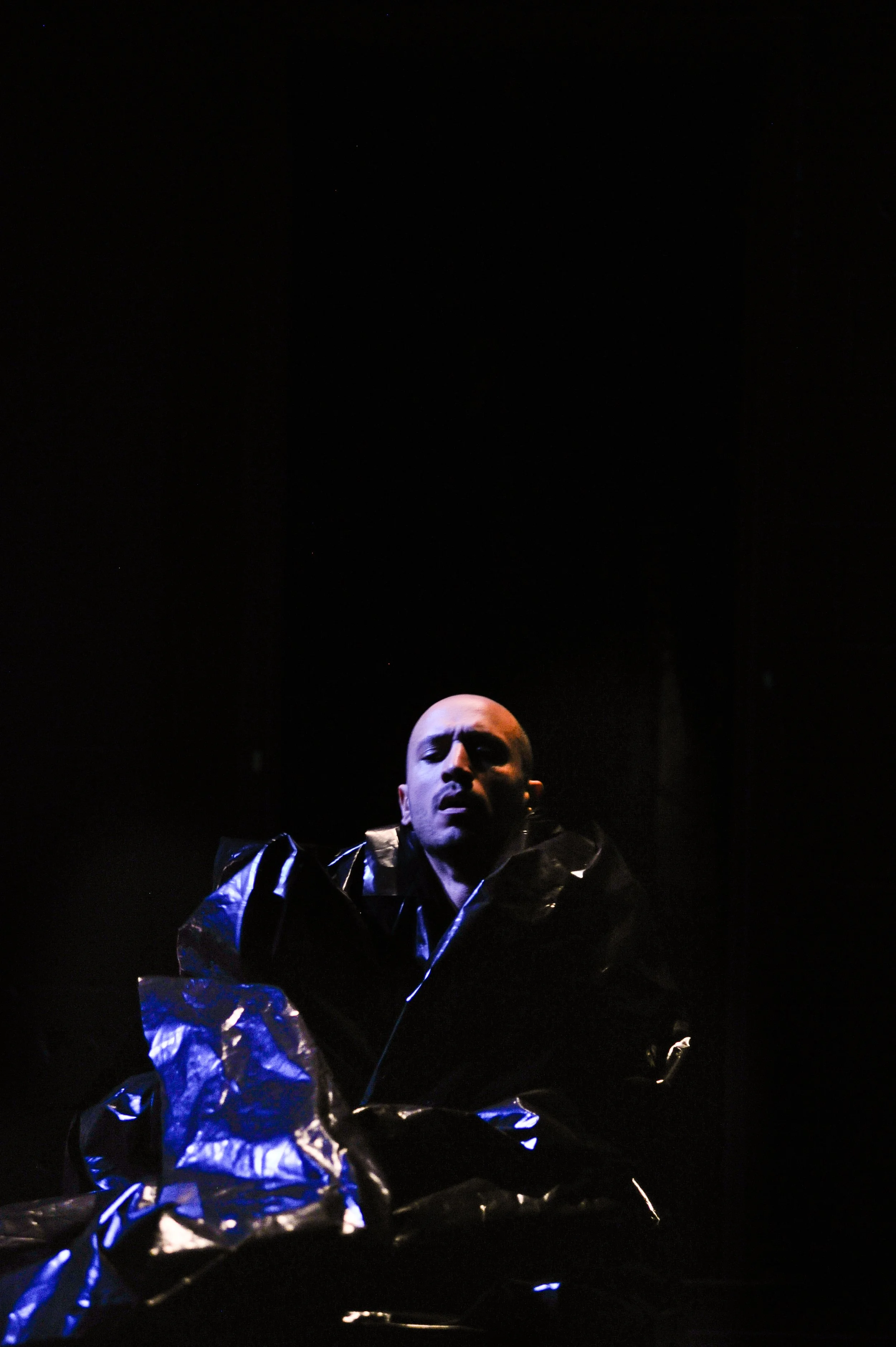 A person with short hair dressed in a shiny, reflective black jacket, looking downward against a dark background with a faint starry sky.