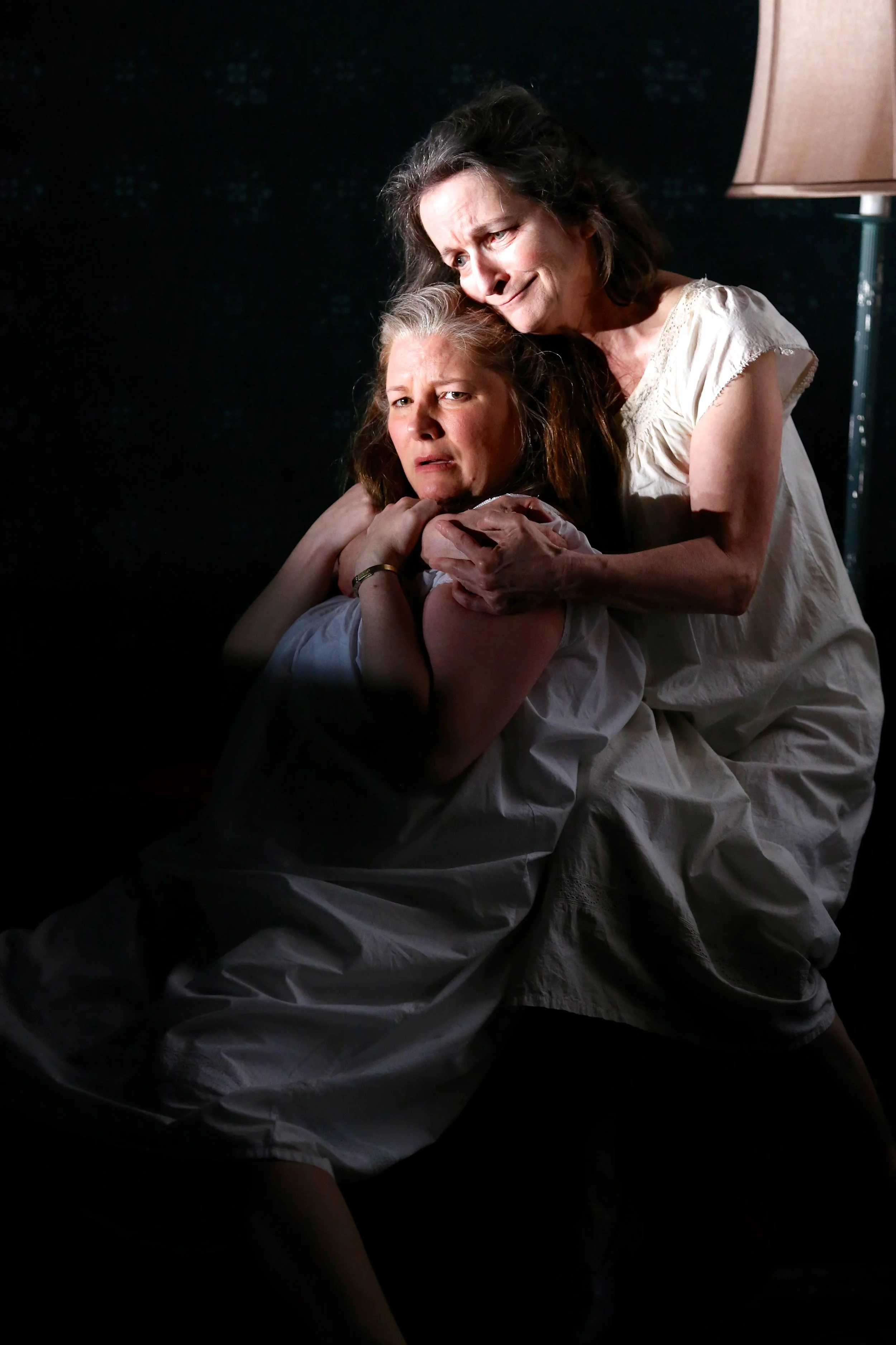 Two women, one middle-aged and the other younger, sharing an emotional moment in a dimly lit room. The older woman embraces the younger woman, offering comfort.