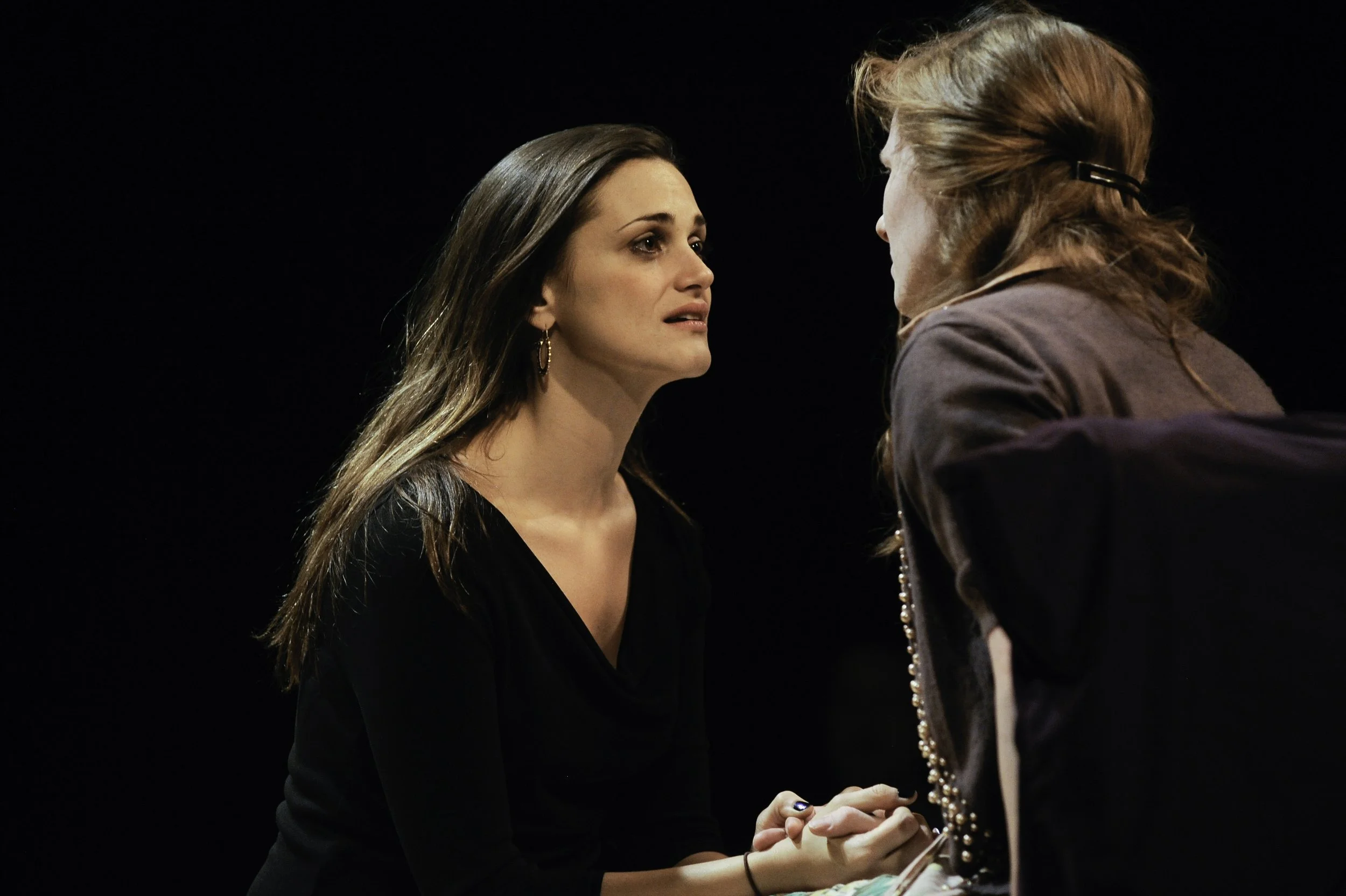 Two women face each other closely, holding hands, on a dark stage. The woman on the left has long brown hair, earrings, and a black top. The woman on the right has reddish hair pulled back with a hair clip and wears a brown jacket. They appear to be engaged in an emotional moment.