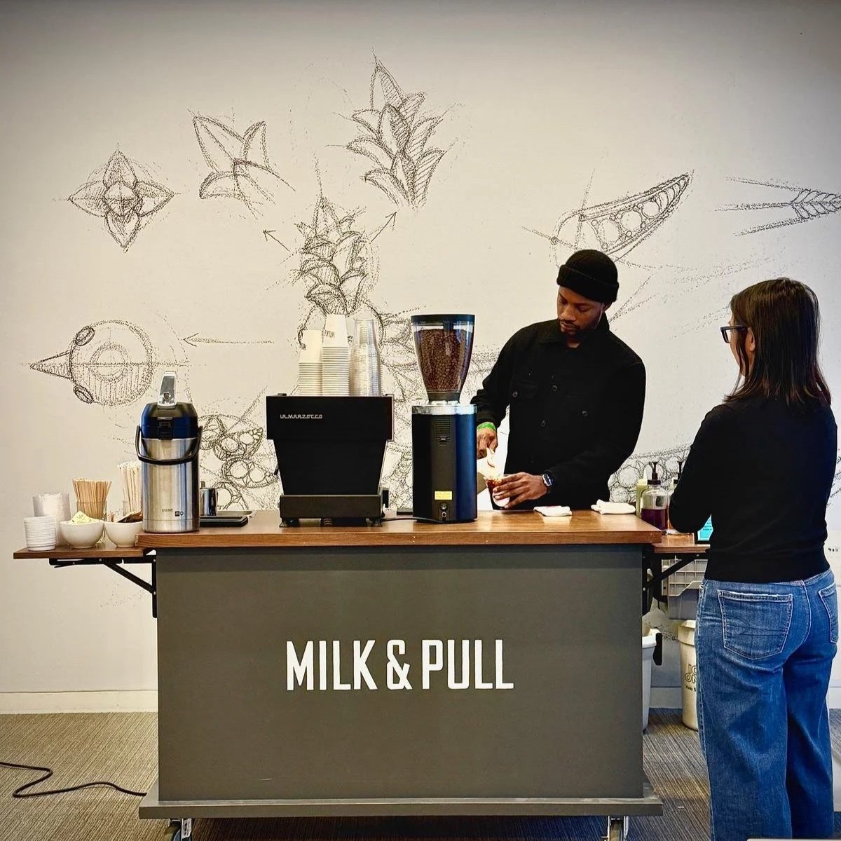 A barista making coffee at a coffee cart labeled "Milk & Pull" at a brand activation event. There is a woman standing in front of the counter. The background has a wall with sketches of coffee-related drawings. Coffee Cart  Event catering