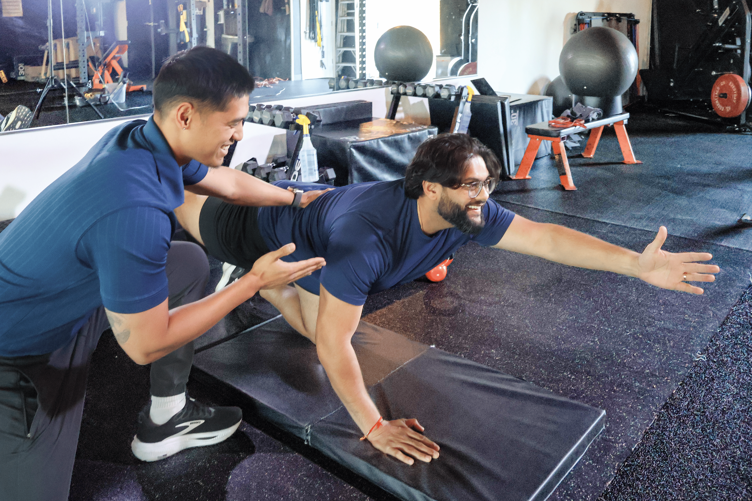 Physical therapist assisting patient performing a plank exercise. The background features gym equipment such as dumbbells, medicine balls, and weight plates.