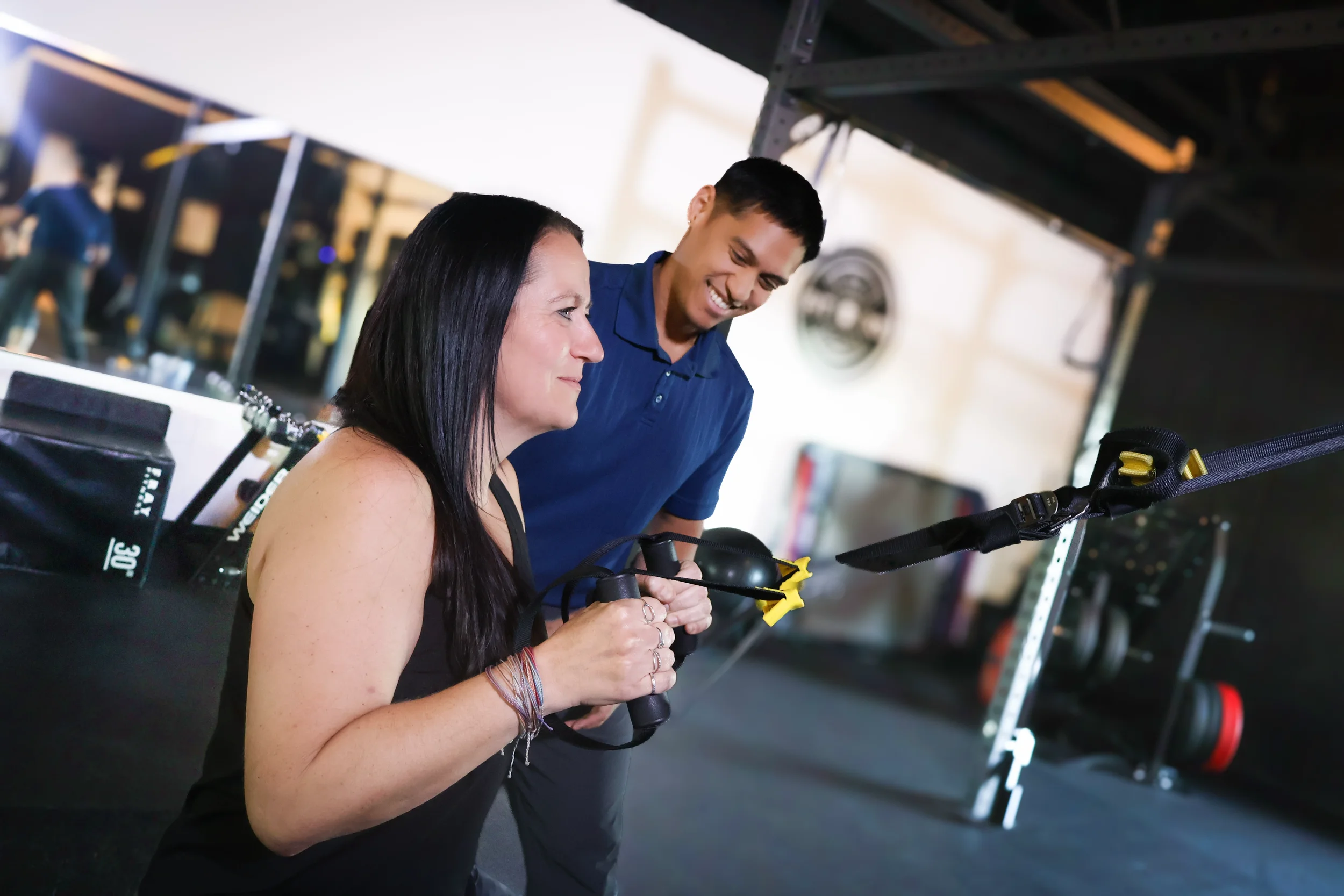 A woman and a man smiling and looking at a piece of equipment in a gym or fitness center.