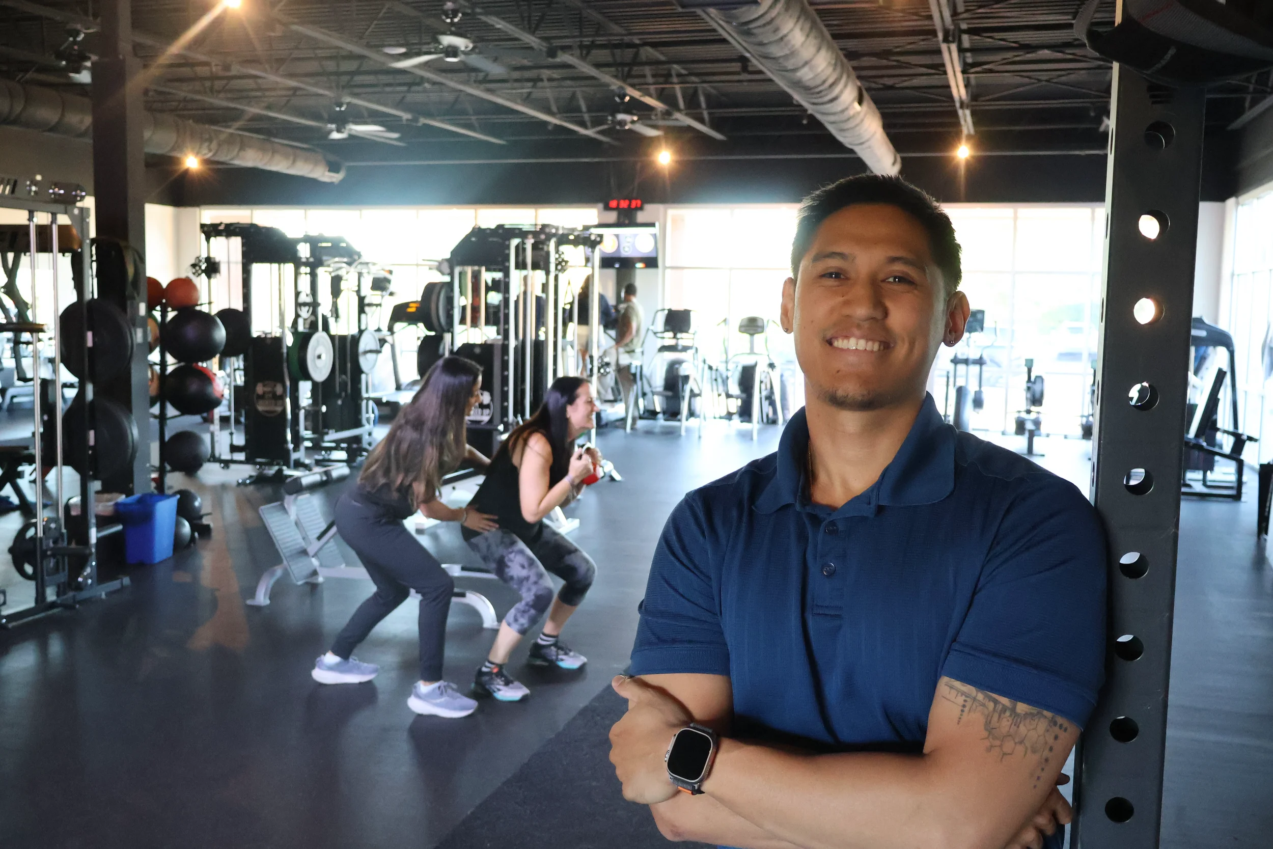 A man smiling inside a gym, taking a selfie in front of two women exercising on a bench. The gym has weightlifting equipment and large windows letting in natural light.