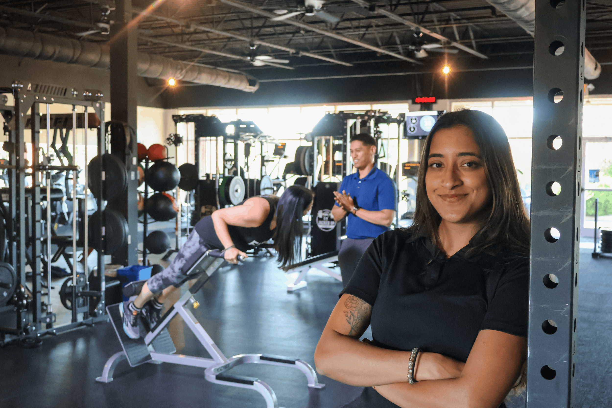 A woman with long dark hair and tattoos on her arm smiling at the camera inside a gym. In the background, people are exercising with weights and gym equipment, with sunlight coming through large windows.