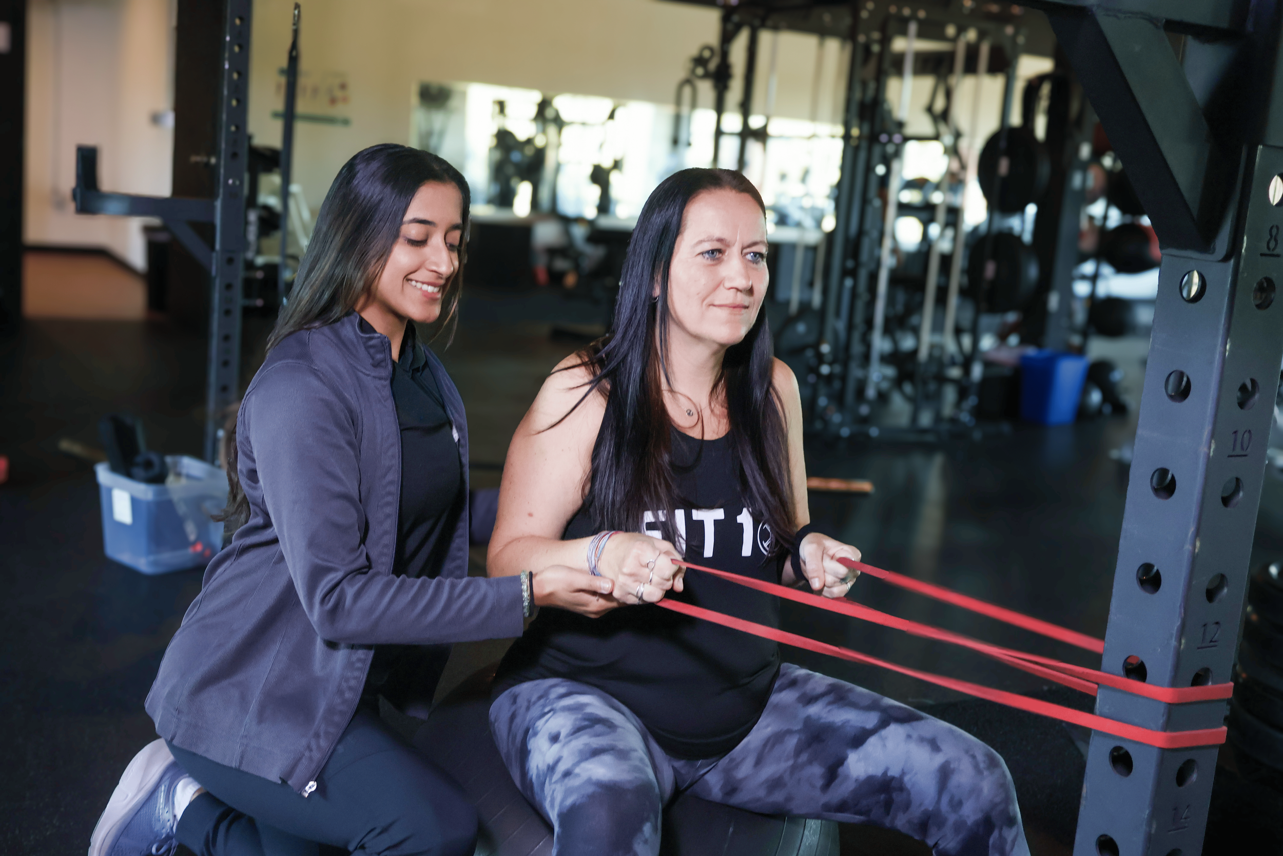 Physical therapist assisting a woman with resistance band exercises in a gym with workout equipment in the background.