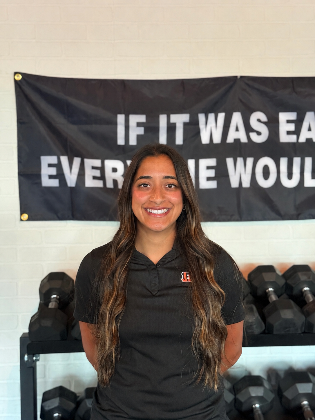 A young woman with long wavy brown hair and a black sports shirt standing in front of a black banner with the partial text 'IF IT WAS EASY EVERYONE WOULD DO IT'. She is smiling and standing with crossed arms in a gym with black dumbbells on racks in the background.