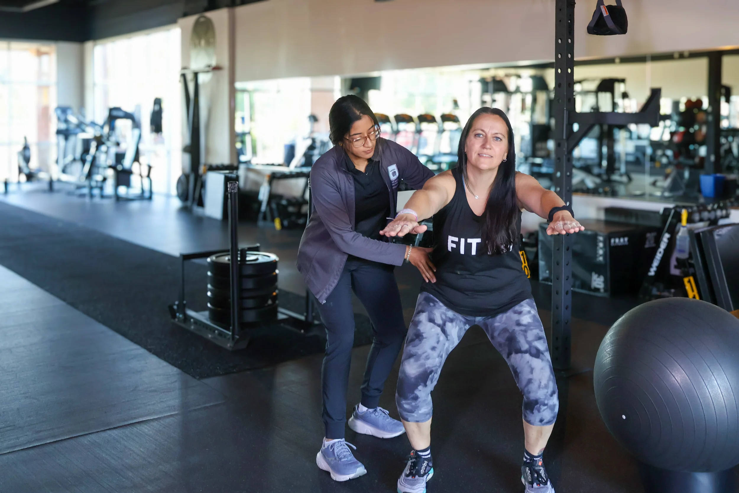 A woman performs a squat with a large medicine ball in a gym, assisted by a trainer.