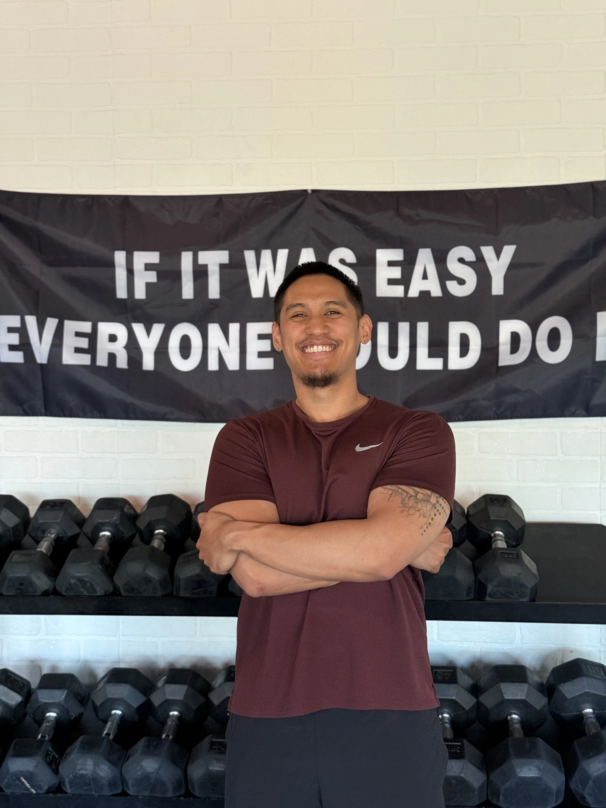 A smiling man with crossed arms standing in front of a wall with a black banner. The banner has text that reads, 'If it was easy everyone could do it,' with a row of black dumbbells underneath.