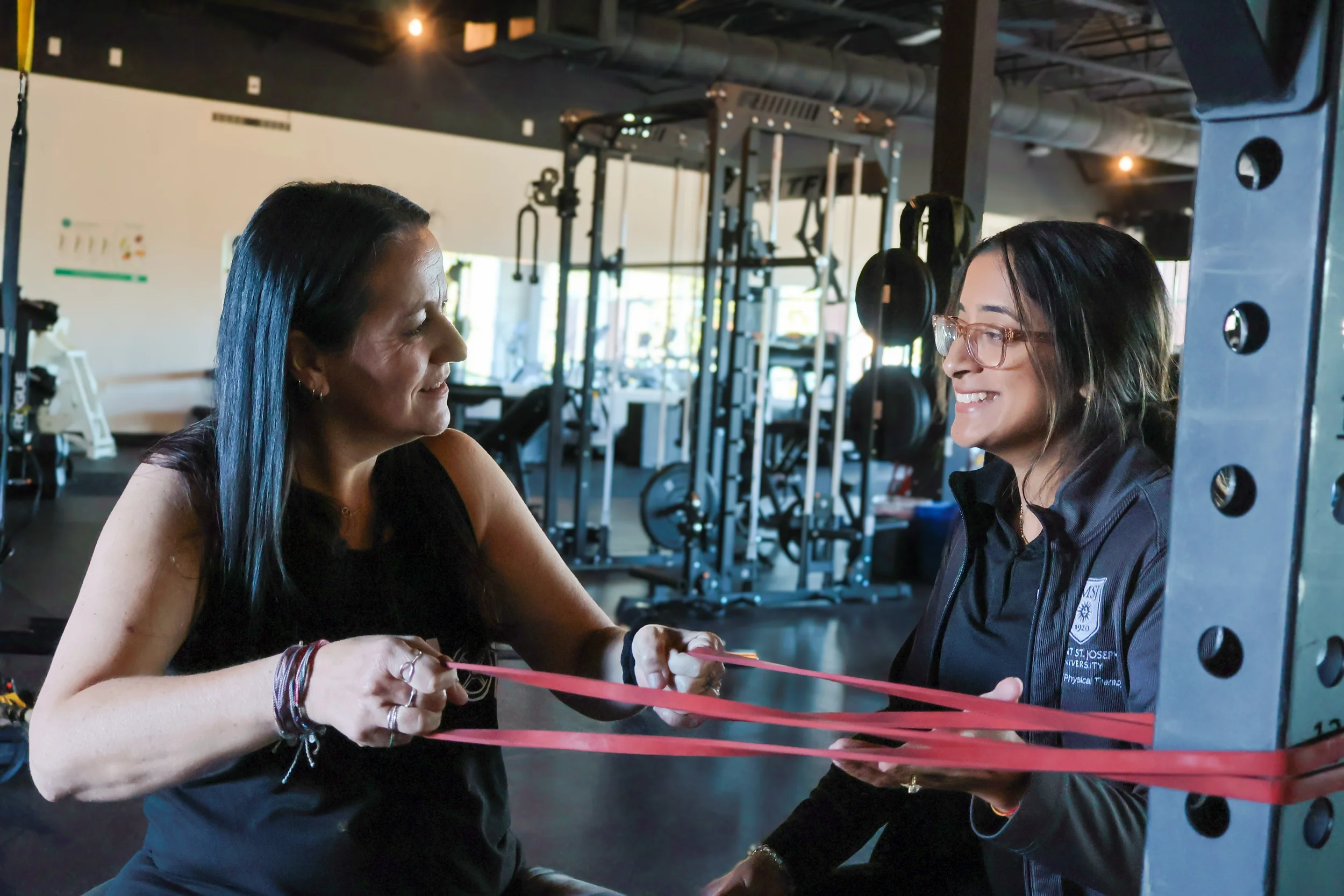 Two women smiling and tying red ribbons together in a gym or fitness studio with exercise equipment visible in the background.